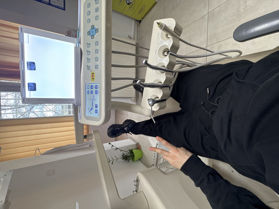 The image depicts an interior scene within a dental office, identified as likely located in Sofia, Bulgaria, based on the "AOL" branding on the dental equipment, which corresponds to a Bulgarian dental manufacturer.
A person is seen from a first-person perspective, lying in a white dental examination chair. They are dressed in a black hooded sweatshirt and black sweatpants, with black sneakers visible on their feet. Their left hand is raised into the frame, forming a two-finger "peace" or "victory" sign.
To the right of the person, a white dental instrument console is prominently displayed. It features a digital control panel with various settings and a diagram, and the "AOL" logo is visible. Attached to the console is a tray holding multiple dental tools, including an air-water syringe, a high-speed handpiece, a low-speed handpiece, and another tool possibly an ultrasonic scaler or curing light, all connected by grey hoses. Above the console, an X-ray film viewer (illuminator) displays a bright white screen with two dark rectangular clips.
The background shows a white wall with a window fitted with brown vertical blinds, through which bare tree branches are visible, suggesting an exterior environment. A small green potted plant sits on the floor against the wall. A white lab coat hangs on a hook on the left wall. The floor is covered with light-colored tiles. A yellow or lime green drawer unit is partially visible on the far right, with a calendar or document holder hanging on the adjacent wall. The overall environment appears sterile and well-lit.