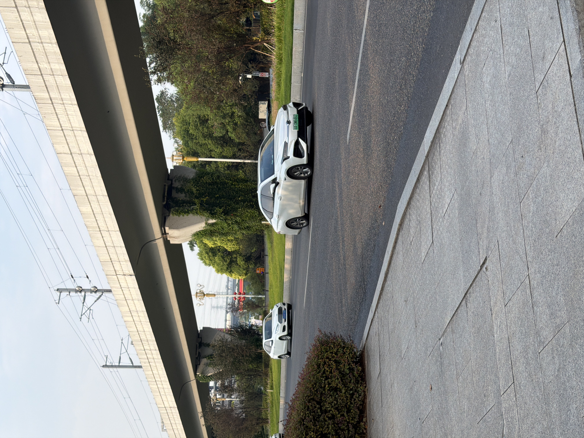 A white sedan with a green license plate, "A DW6462," drives on a paved road in Jiangjin, China. Another white sedan with a blue license plate is visible behind it. The road is bordered by a grey sidewalk on the left and a grassy median on the right. An elevated concrete structure, likely an overpass or bridge, spans the scene, supported by pillars. Overhead, power lines are suspended. The sky is clear and bright, suggesting daylight. Trees and some buildings are visible in the background.