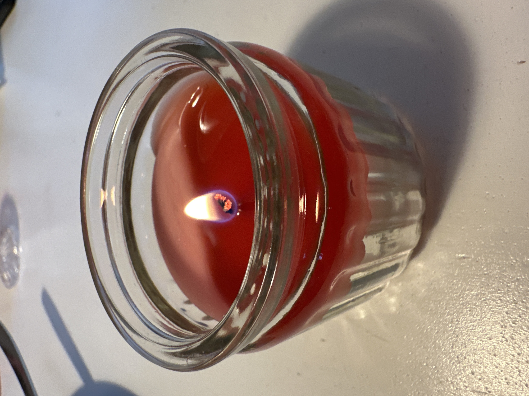 A lit red candle in a glass jar sits on a white surface. The candle has a visible flame and melted wax is visible in the jar. The jar has a ribbed texture along its sides. A shadow of the jar and candle is cast on the surface to the left.