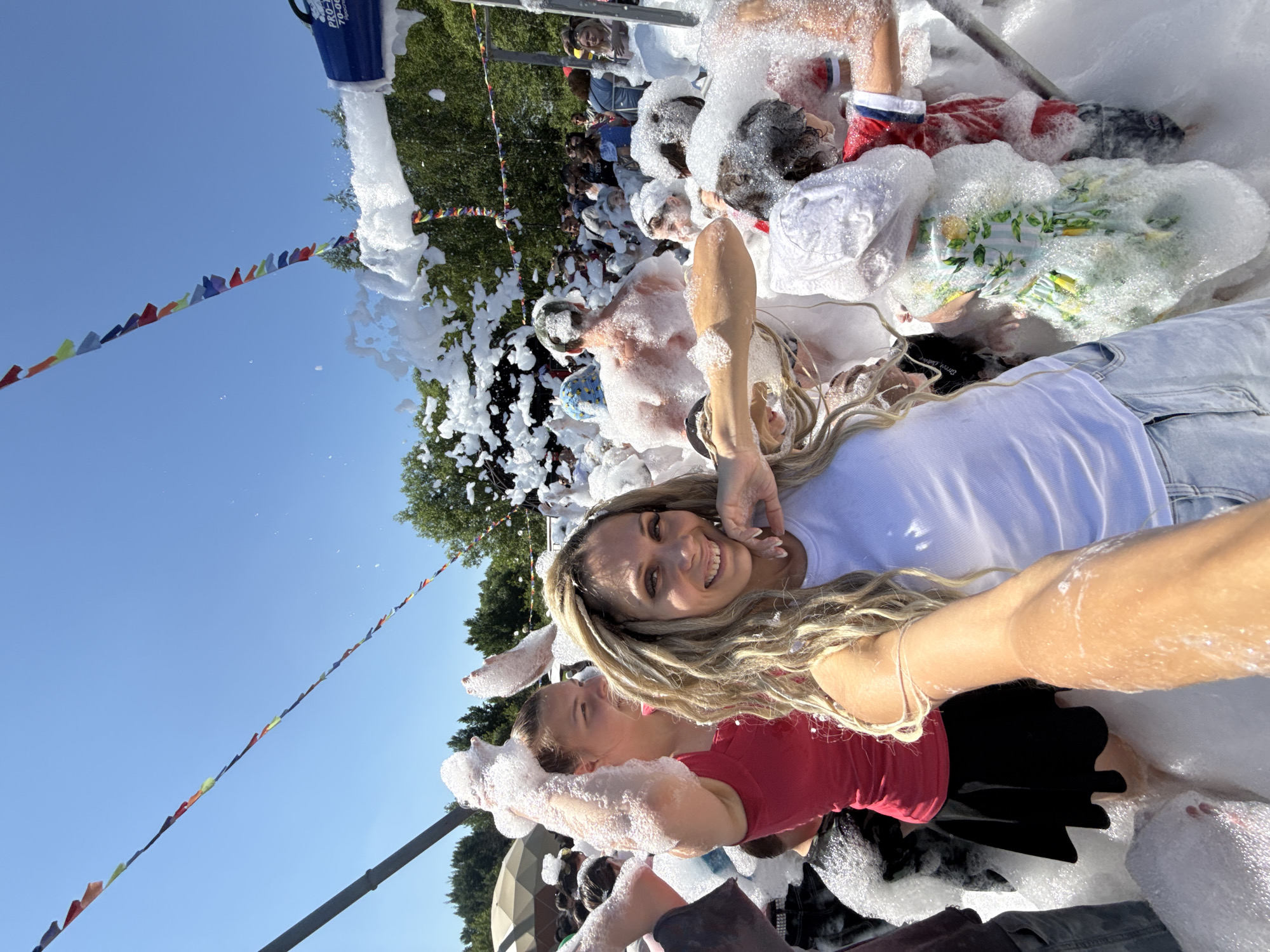 The image depicts a crowded outdoor foam party, possibly in the city of Шебунино, Russia, under a clear blue sky. Several individuals, primarily young adults and children, are immersed in a large amount of white foam. In the foreground, a woman with blonde dreadlocks is taking a selfie, smiling broadly. To her left, another woman in a red top is visible, with her hands raised and covered in foam. The background shows more people enjoying the foam party, with trees and what appears to be a tent structure in the distance. Colorful triangular flags are strung across the scene, adding to the festive atmosphere. A foam machine is partially visible on the right side of the image, actively producing more foam. The overall scene is one of playful and energetic celebration.