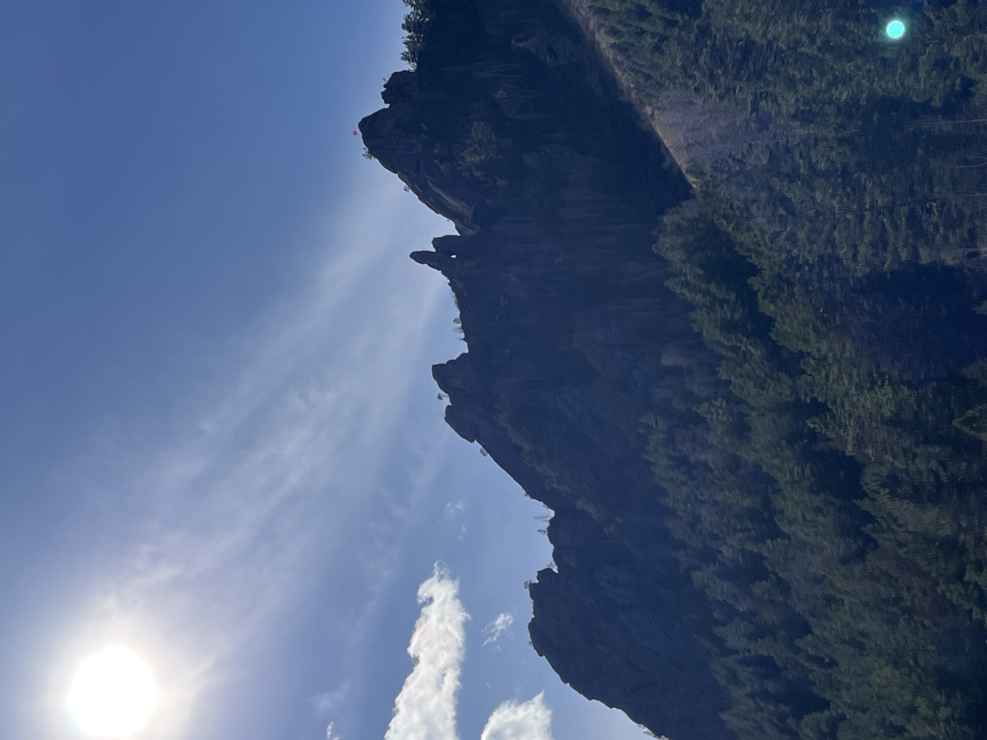 The image shows a rocky cliff face with trees along its side, set against a blue sky with some clouds and the sun visible in the lower left corner. There is a red object atop the cliff. The location is likely in the Krasnoyarsk region of Russia, given the mention of Krasnoyarsk. The scene is outdoors during daylight.
