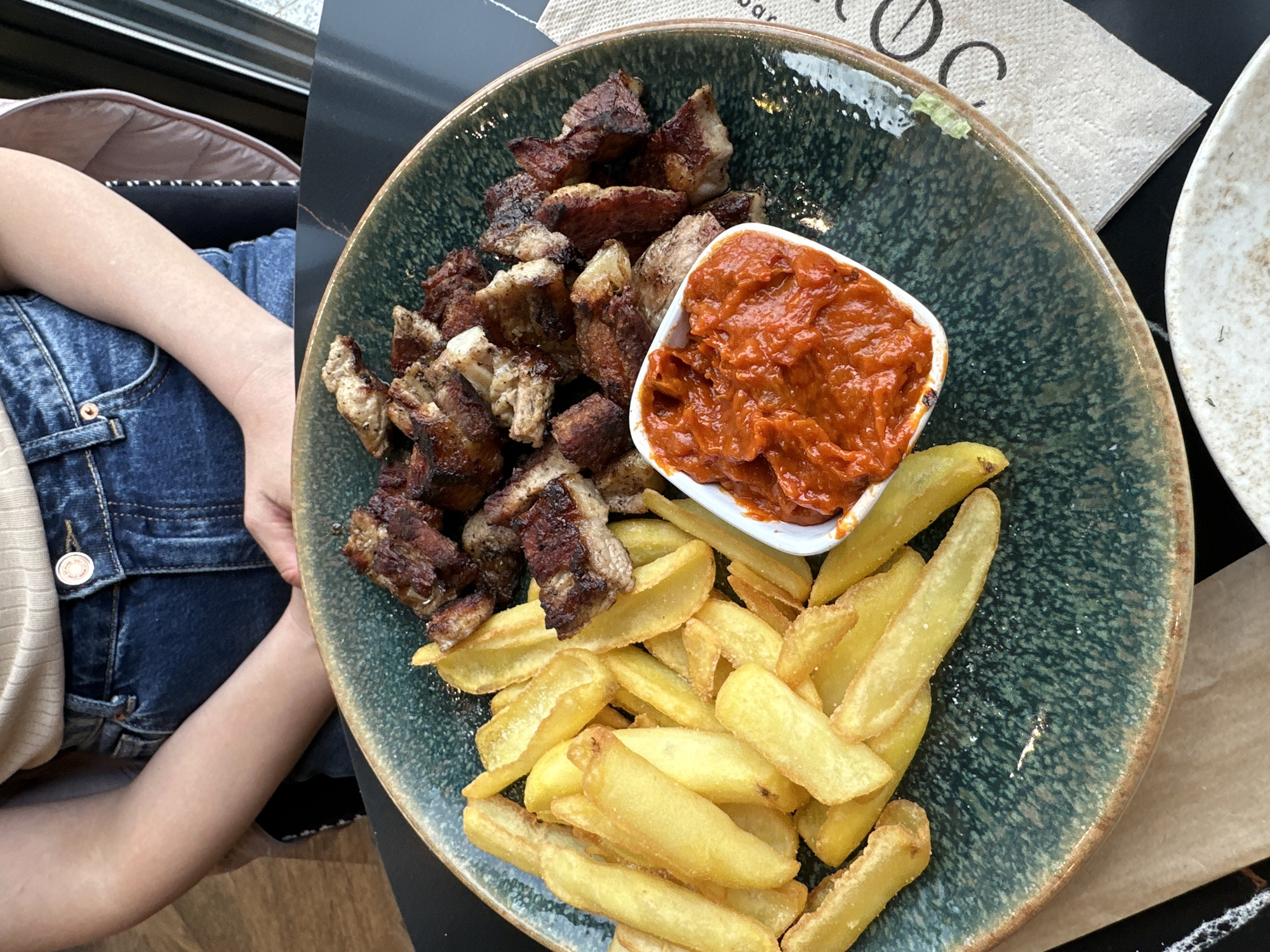 A top-down, eye-level shot captures a meal of fried pork cubes and French fries with a red pepper sauce, served on a green speckled plate with a brown rim. A white square dish holds the sauce. The plate sits on a black and brown wooden surface, and a second white plate is partially visible to the right. A person, wearing blue denim jeans, is seated to the left of the plate. The scene is indoors, next to a window, potentially in a restaurant. The city and country location of Montana, Bulgaria, is provided, but cannot be verified from the image alone.