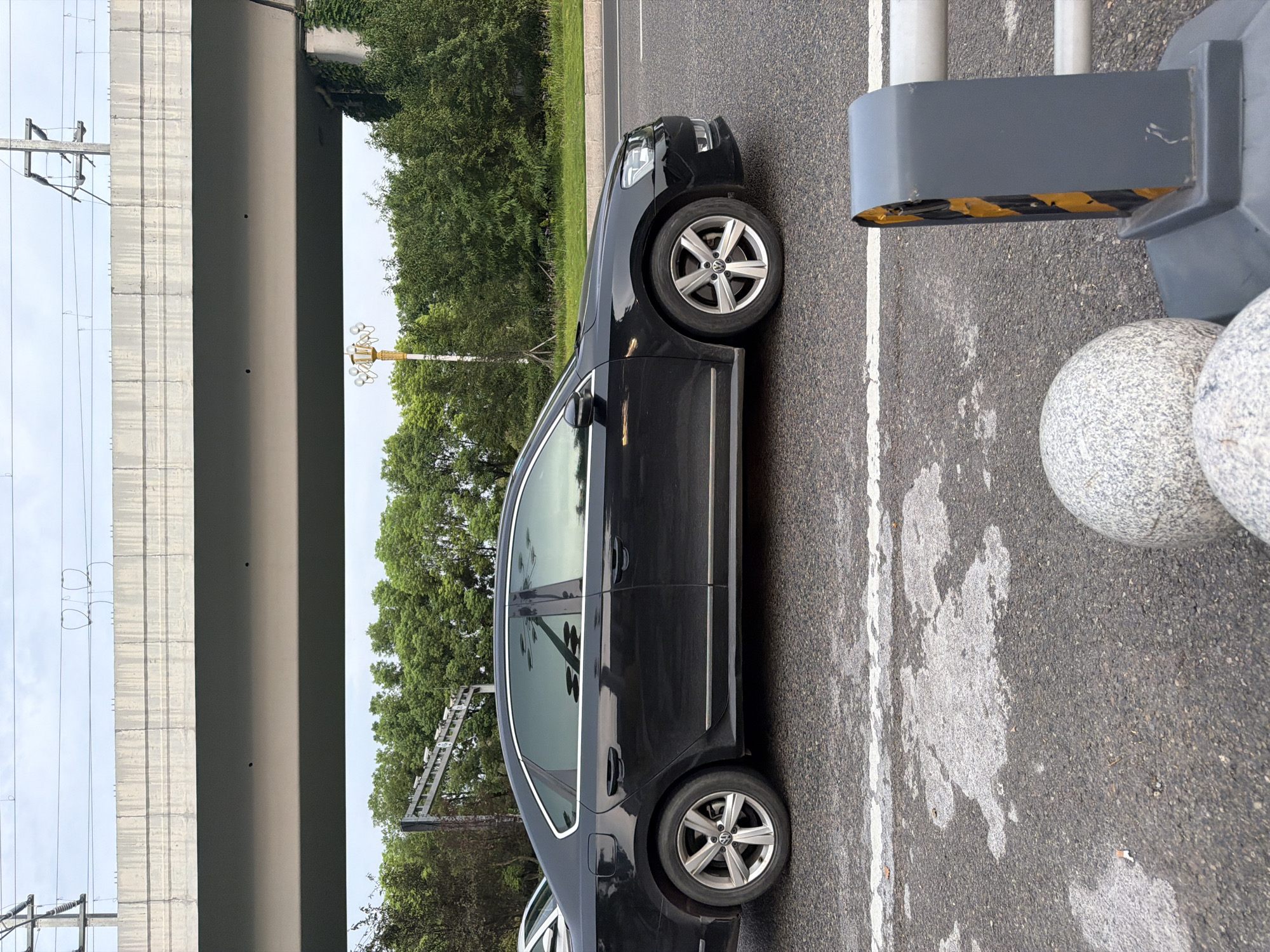 The image shows a black Volkswagen sedan driving on a road in Jiangjin, China. The car is traveling from left to right. In the background, there is an elevated highway or overpass with overhead power lines. Trees and greenery are visible beyond the road. In the foreground, there are road markings and traffic control devices, including a barrier with yellow and black stripes and decorative spherical objects. The sky is overcast.