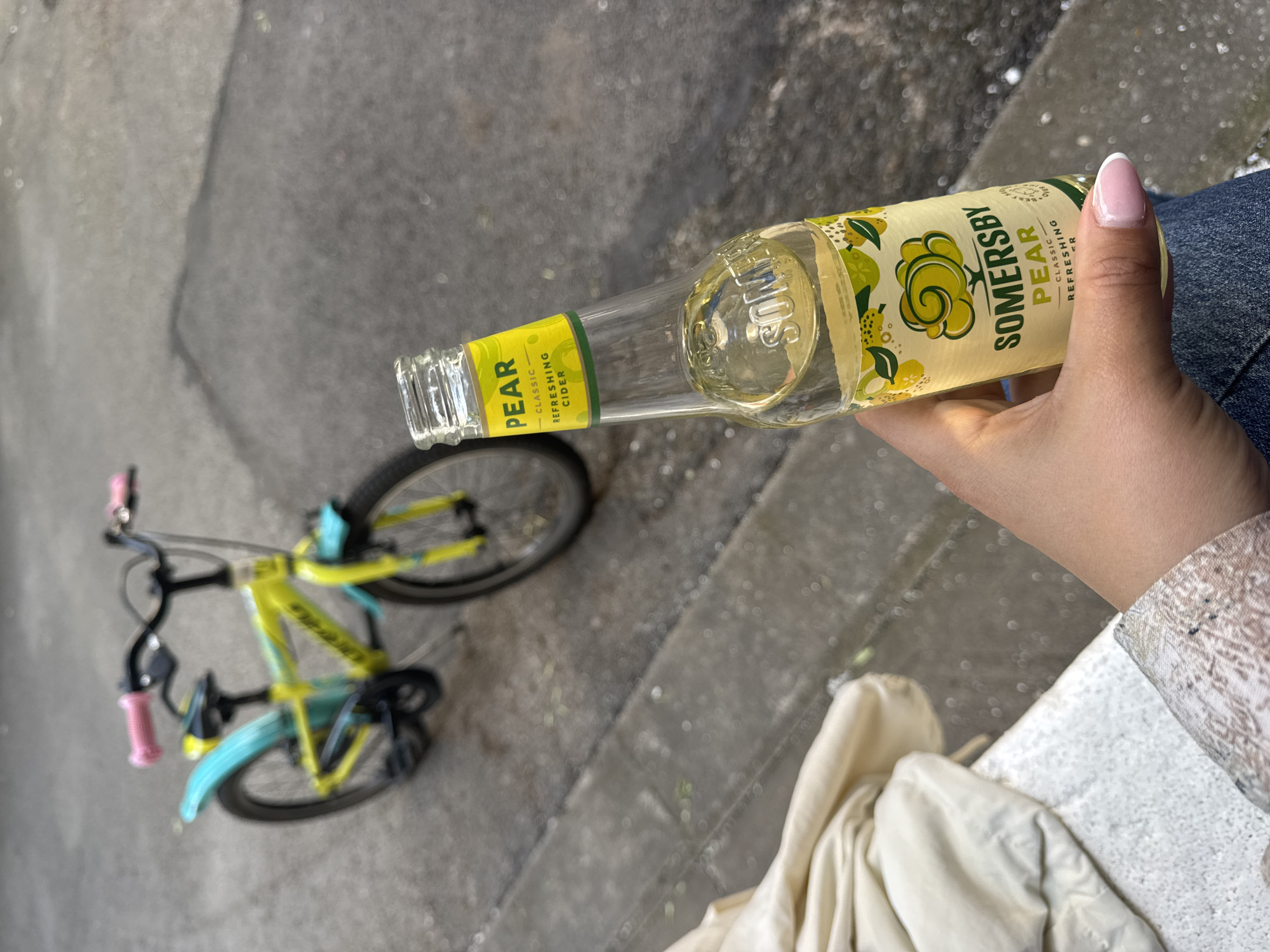 A hand is holding a partially consumed bottle of Somersby Pear cider at an angle in an outdoor setting, likely a street. The bottle's label is clearly visible, indicating the brand and flavor of the beverage. To the left, a bicycle with a distinctive color scheme (yellow frame, blue fenders, and pink handlebar grips) is parked on the sidewalk. The ground is paved and appears to be made of concrete. The scene is likely in an urban area. The geographical location of Sofia, Bulgaria, cannot be determined from this image.