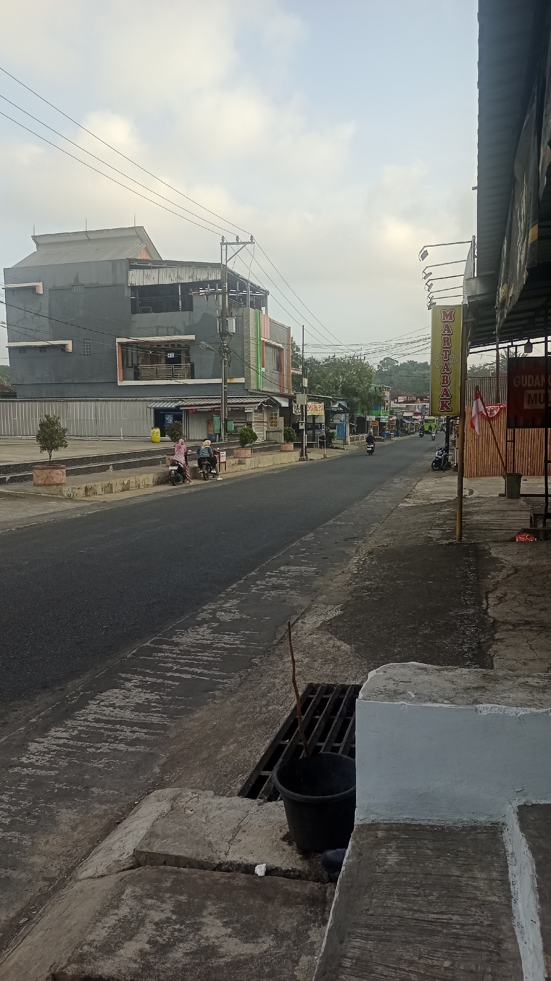This is a street-level view of a road in Pakemitan, Indonesia, under an overcast sky. The road is paved and appears wet in some areas. On the left side of the road is a raised concrete walkway with a building behind it. The building has a modern design with dark grey and orange sections, and multiple windows. Several potted trees are placed along the walkway. A few people on motorcycles are visible, some parked and others riding on the road.
On the right side of the road, there is a covered awning extending from a building. A yellow sign with red lettering that reads "MARTABAK" is prominent. Another sign above it reads "MASTER" in smaller letters. Further down the street, more buildings and shops can be seen, with trees and foliage in the background. A red and white flag is displayed near the martabak sign. A black plastic bucket with a stick protruding from it is placed next to a metal grate on the sidewalk in the foreground. The overall scene depicts a commercial street, possibly in the morning or late afternoon.