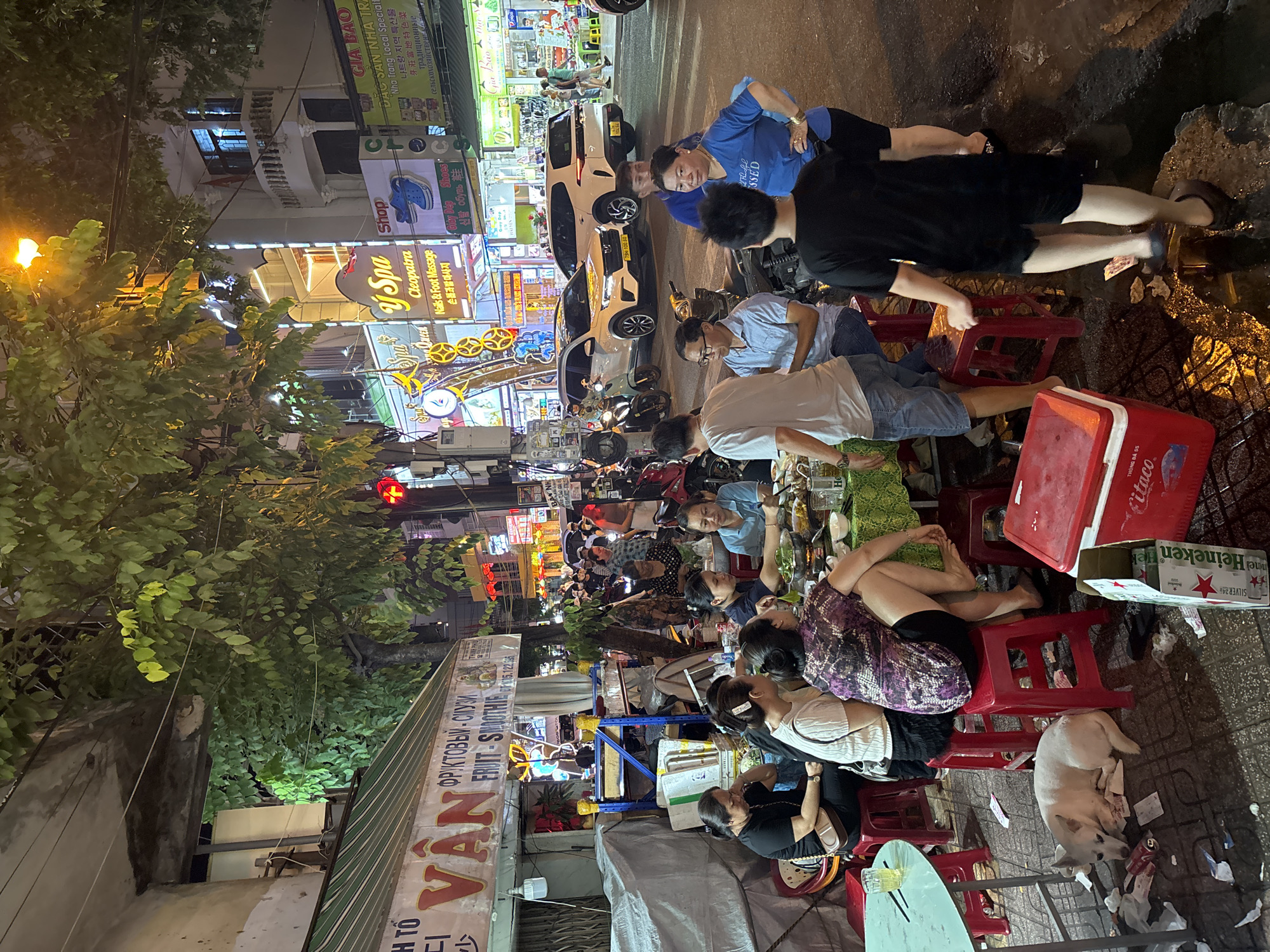 This media file depicts a vibrant street scene at night in Nha Trang, Vietnam. The main subjects are groups of people gathered at outdoor tables, seemingly enjoying a meal or drinks. The setting is a bustling street with numerous neon signs advertising shops and services. Visible text includes "VAN", "FRUIT-SMOOTHIE", "Spa Shop", "Cleopatra Nails & Foot Massage", "Giày Dép Shoes", "GIA BAO", and "Heineken". The time of day is clearly night, indicated by the artificial lighting from the signs and the ambient darkness. The atmosphere appears lively and social, with people engaged in conversation and activity. There are also several parked motorbikes and cars, suggesting a busy urban environment. A white dog is resting on the ground near some of the tables. The overall impression is one of a popular local gathering spot in a lively Vietnamese city.