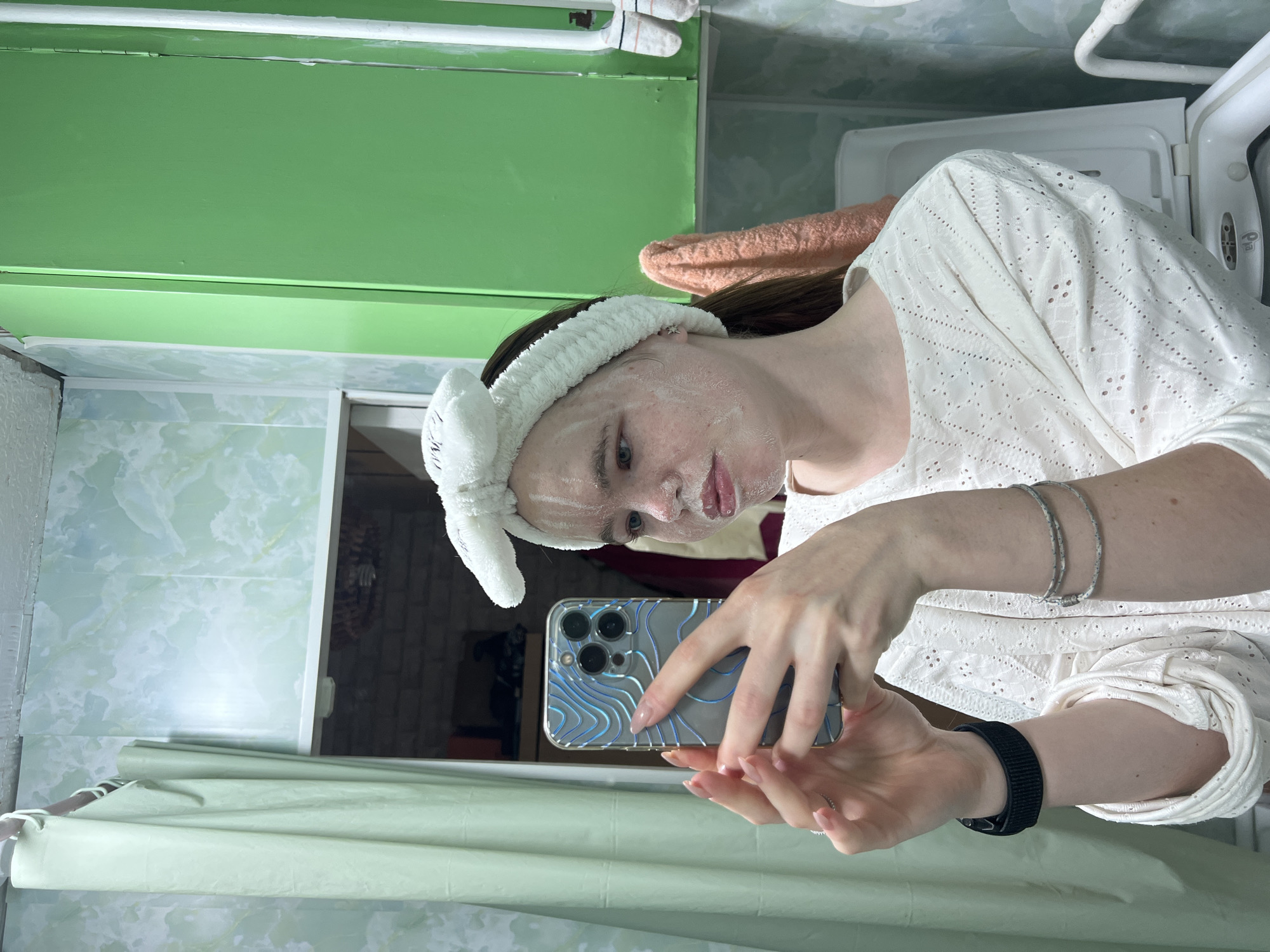 A woman takes a selfie in a bathroom. She is wearing a white shirt with eyelet embroidery and a white headband with bunny ears. She has a face mask on. Her left arm is adorned with bracelets and a black watch band. She is holding a blue-and-silver iPhone in her hands. The bathroom has green-painted cabinets and green-and-white marble-look tiles. An orange towel is draped on the cabinet above the toilet. A washing machine is visible to the right. Based on the request, this location is in the city of Chaikovsky, Russia.