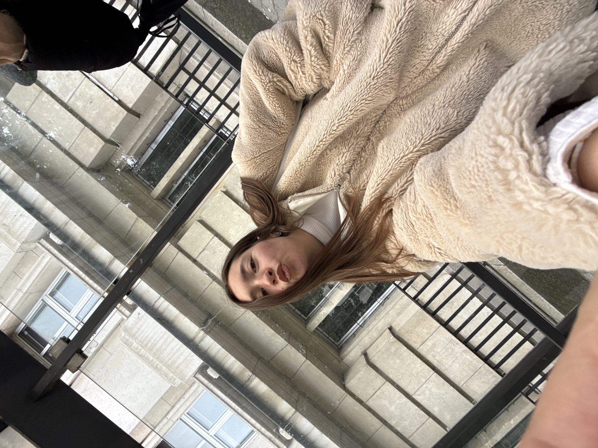 A young woman with brown hair and a beige fleece jacket is positioned in front of a glass floor or surface, through which a building facade is visible. The building appears to be multi-storied with a light-colored, possibly stone or concrete exterior, featuring multiple windows. Black railing frames a portion of the view below. This image was taken at the Largo, an architectual ensemble in Sofia, Bulgaria.