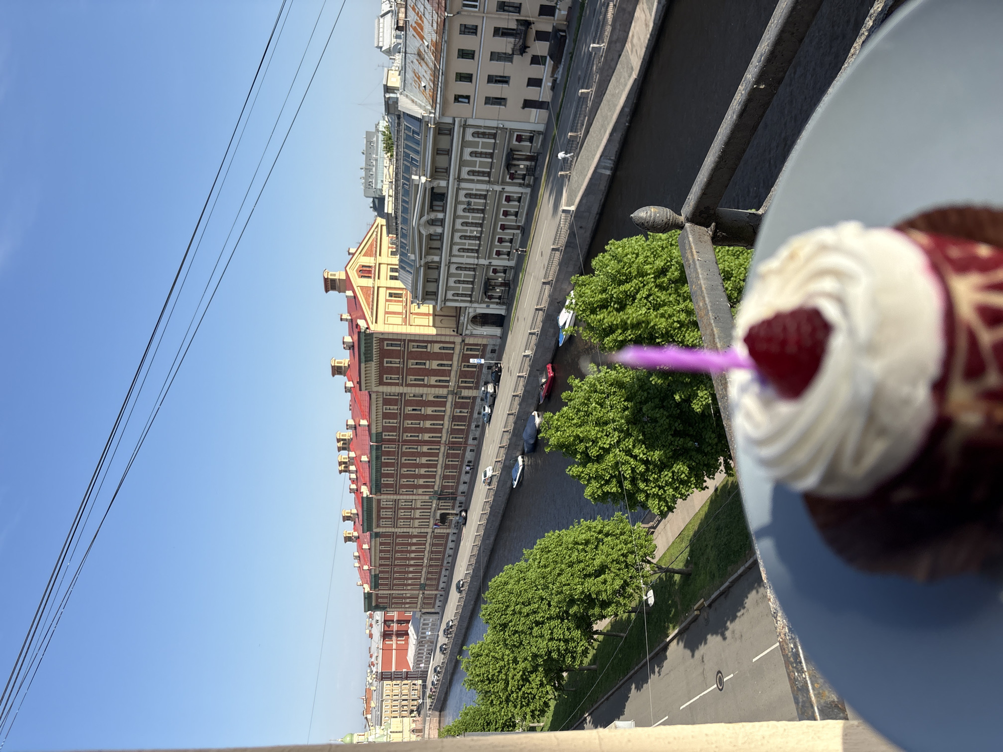 The image depicts a cityscape in Saint Petersburg, Russia, viewed from an elevated position. In the foreground, a cupcake with white frosting and a raspberry is placed on a plate, positioned near a wrought-iron railing. Beyond the railing, a canal runs parallel to a street lined with buildings of varying heights and architectural styles, most of which are constructed in the style of traditional European architecture. There are trees and vegetation visible along the canal. The sky is clear and blue, and electrical wires stretch across the upper left corner of the frame. People and vehicles are on the street next to the buildings.