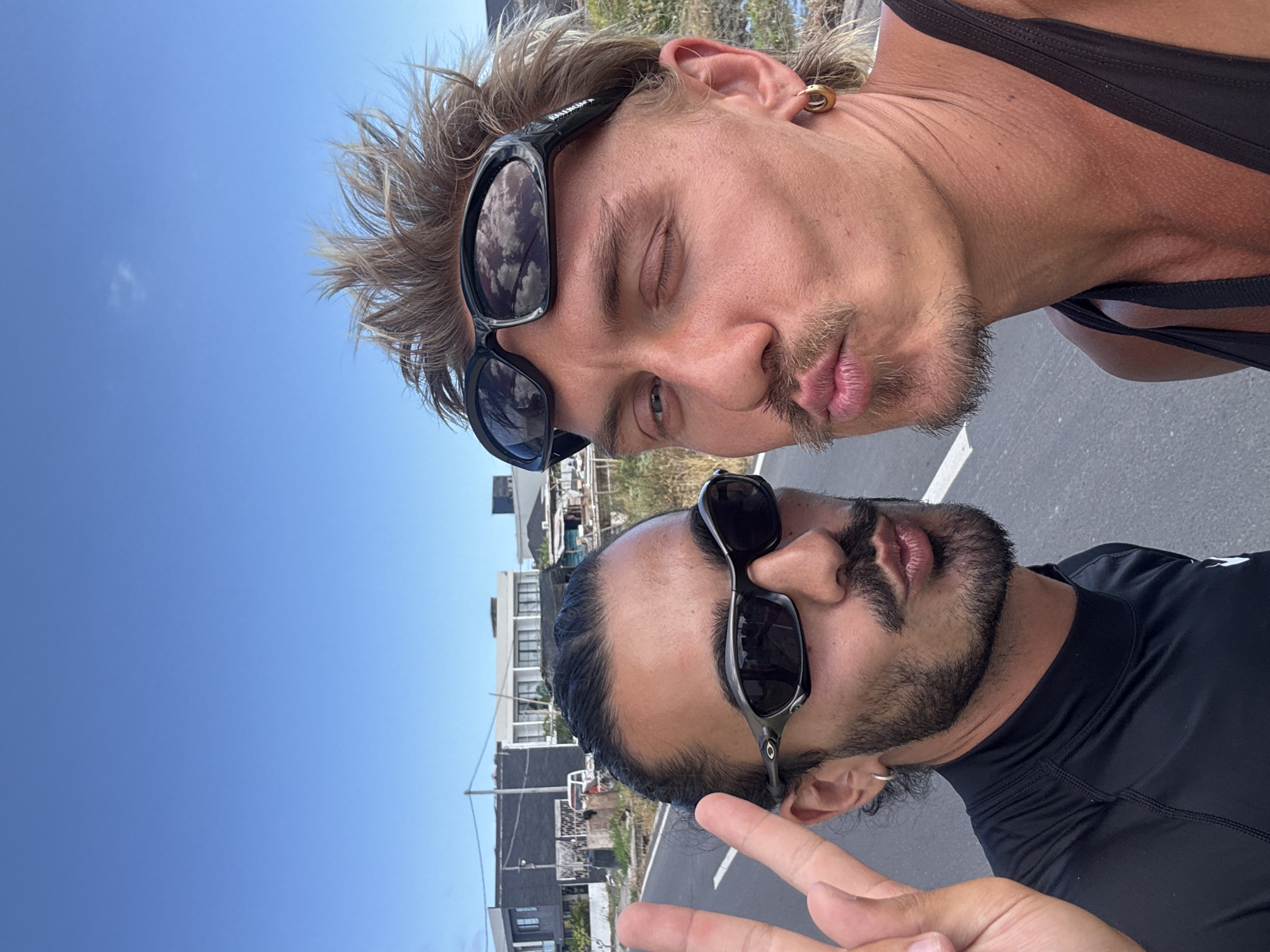 Two men pose for a selfie. The first man, positioned at the top of the frame, has blonde hair, sunglasses with the word "Balenciaga" on the side, and an earring. The second man, positioned at the bottom of the frame, has dark hair, a beard, sunglasses, and is holding up two fingers in a peace sign. The background reveals buildings, utility poles, a blue sky and street. The image was most likely taken in Mengwi, Indonesia.
