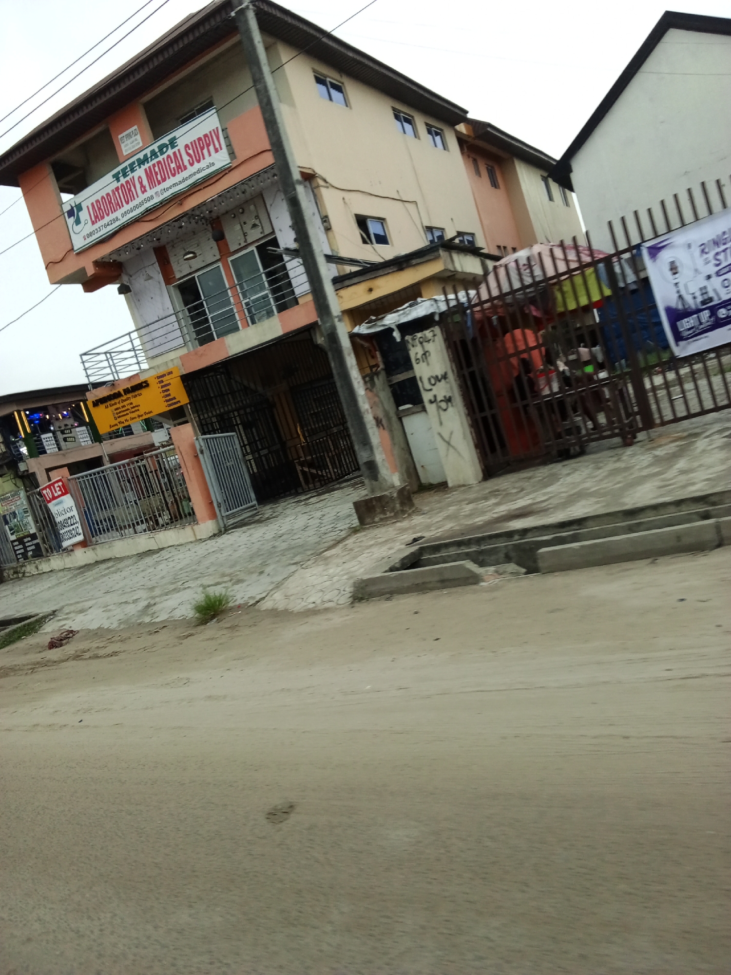 A corner building in a city, possibly Lagos, Nigeria, is captured from a slightly elevated, oblique angle. The building, painted in shades of peach and cream, has multiple stories with windows and balconies. A prominent banner on the peach section reads "TEEMADE LABORATORY & MEDICAL SUPPLY." Adjacent to this, a yellow sign displays "AFRIKA FABRICS All Kinds of Quality Fabrics." A fenced-off area with iron bars is situated in front of the building, containing some stored items and a metal gate. To the right, another building, a lighter color, has a fence with posters. The ground in the foreground is a dusty, unpaved road or sidewalk, with a concrete drainage channel. The sky above is overcast.