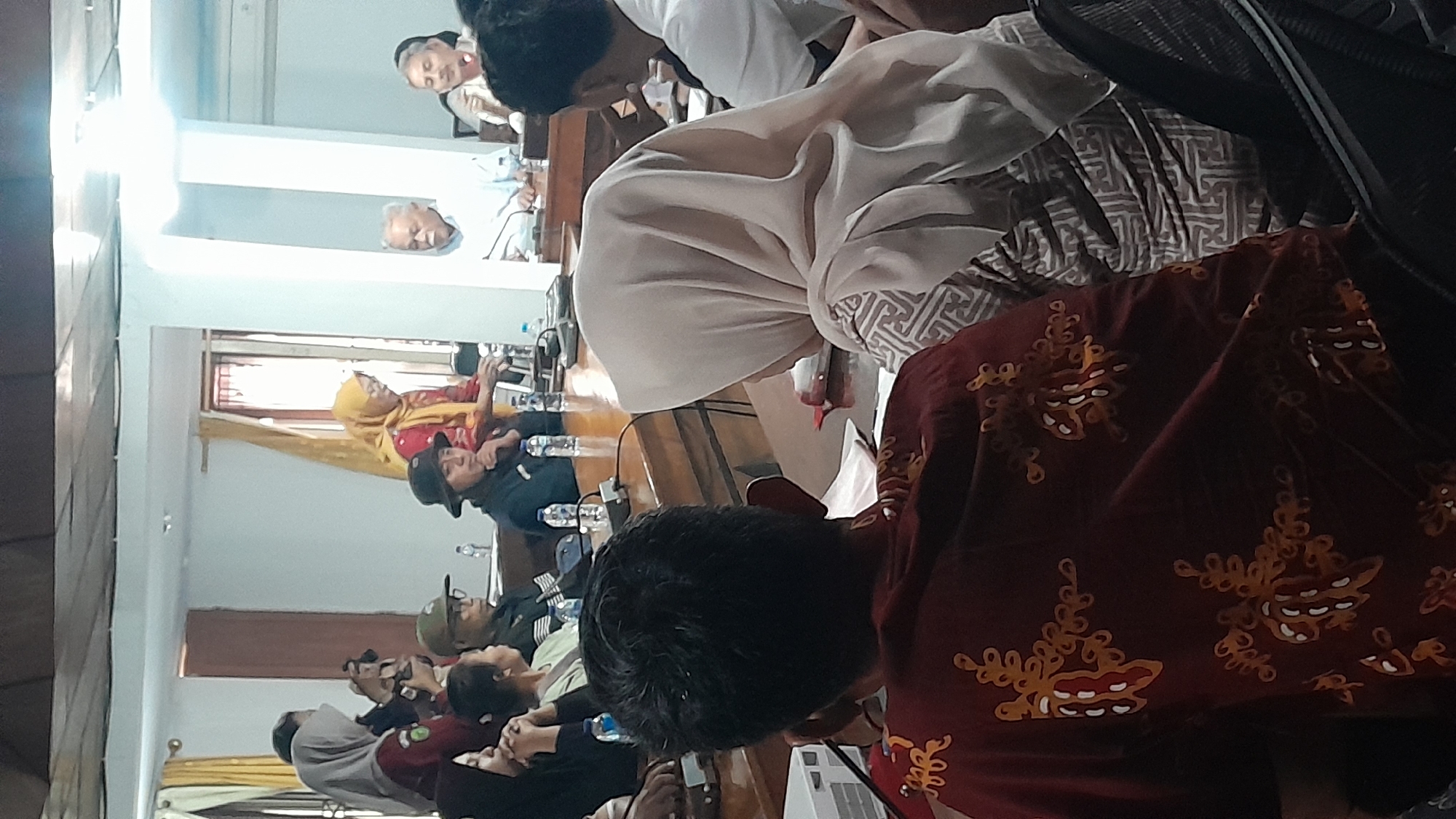 The image depicts an indoor gathering of approximately ten people, seated around a wooden table, possibly for a meeting or conference. The setting appears to be a room with white walls and cream-colored curtains. In the foreground, the back of a person wearing a maroon shirt with an intricate yellow and white pattern is visible. To their right, another person wearing a light-colored headscarf and a patterned dress is seated. Several other individuals are visible in the mid-ground and background, some wearing headscarves, others hats, and one person in a white shirt. Objects on the table include water bottles, microphones, and papers. One person, a woman in a yellow headscarf and red top, is standing and appears to be speaking or gesturing. The overall atmosphere suggests a formal or semi-formal gathering. The specific location in Blora, Indonesia, cannot be determined from the visual information alone.