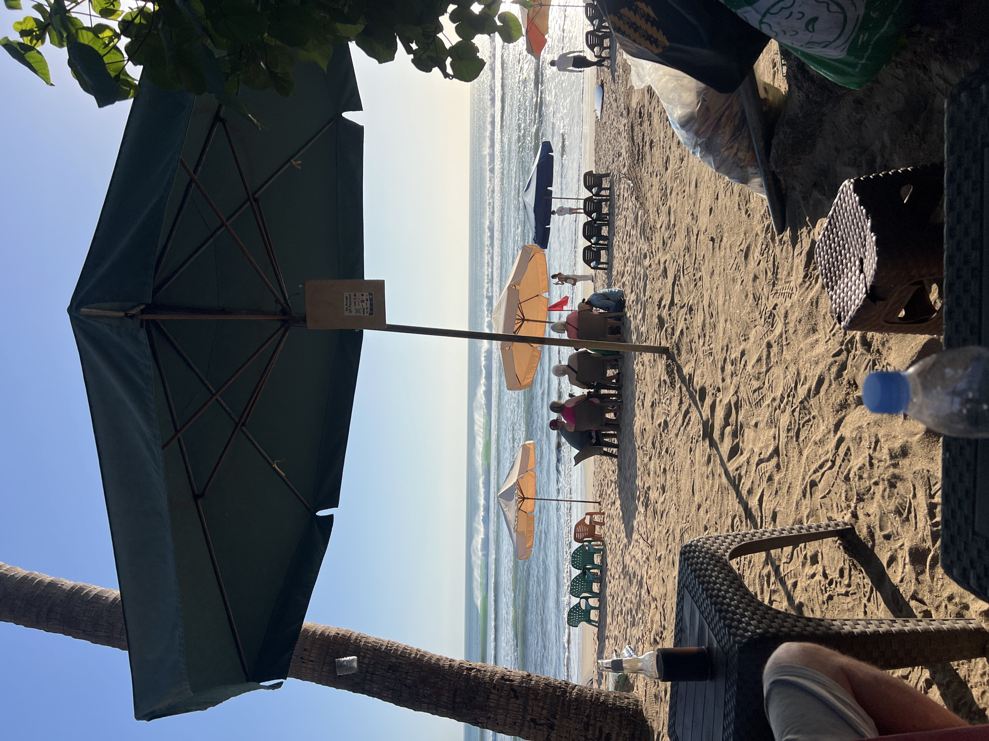 This image depicts a beach scene in Kuta, Indonesia. In the foreground, a person's legs and arm are visible, suggesting they are seated at a dark, woven table. A dark glass bottle sits on the table. To the right, a small plastic stool and a partially obscured plastic bag filled with items are on the sand.
Beyond the foreground, the beach is populated with several beach umbrellas and seating areas. Two orange umbrellas and one dark blue umbrella are set up, with people seated at tables beneath them. A group of four individuals are seated at a table under one of the orange umbrellas, facing the ocean. Another person is standing near the blue umbrella, and two individuals are walking on the beach further down. A white surfboard is leaning against a nearby beach chair.
The ocean in the background features moderate waves breaking on the shore, indicating good surfing conditions. The sky is clear and blue, suggesting a sunny day. A palm tree is prominent on the left side of the frame, with its trunk partially obscured by a large, dark green umbrella that dominates the upper portion of the image. A sign is hanging from the umbrella pole, partially visible. The sand is a light brown color, with footprints and tire tracks visible.