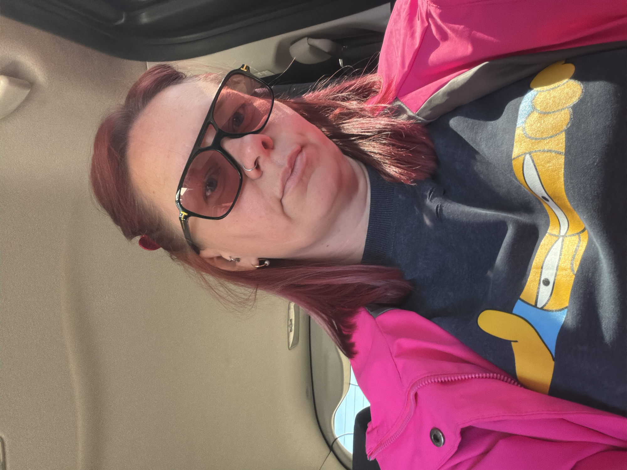 A woman with reddish hair, wearing black-rimmed rose-tinted glasses and a nose ring, sits in what appears to be a car. She is wearing a dark gray sweater with a yellow cartoon figure and a bright pink jacket. The interior of the car is beige. There is no indication of location in the city of Chaikovsky, Russia.