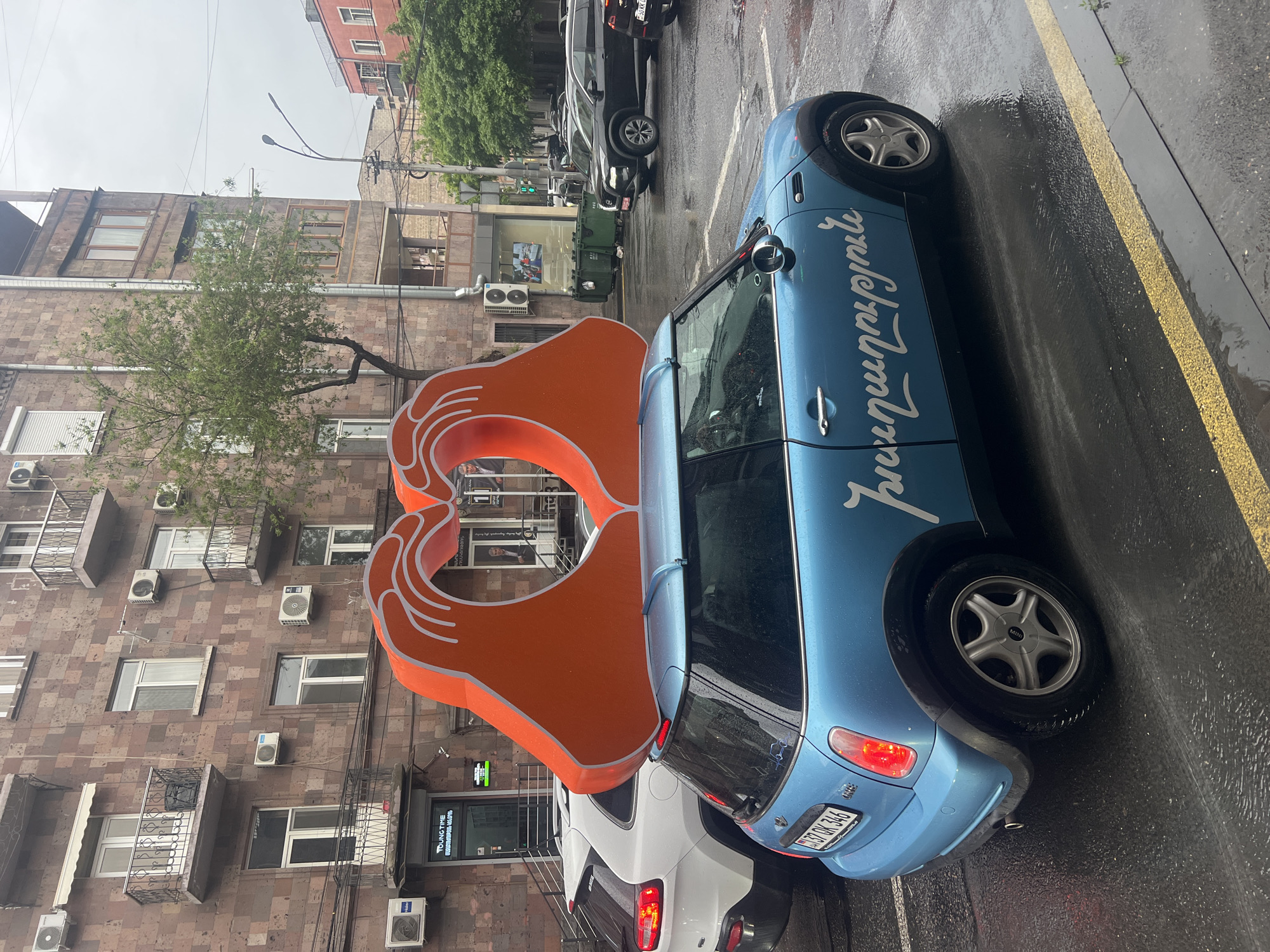 The image shows a street scene in Yerevan, Armenia, on a wet day. A light blue Mini Cooper, adorned with white lettering, is parked on the right side of the frame. One of the car's tires is in contact with the street, and the car's rear is up against another, white car. Behind the Mini Cooper stands a large, orange sculpture depicting two hands forming a heart shape. The sculpture is in front of a building. The building has windows and air conditioning units. In the background, other cars are visible on the street, and taller buildings line the street. The street is wet.