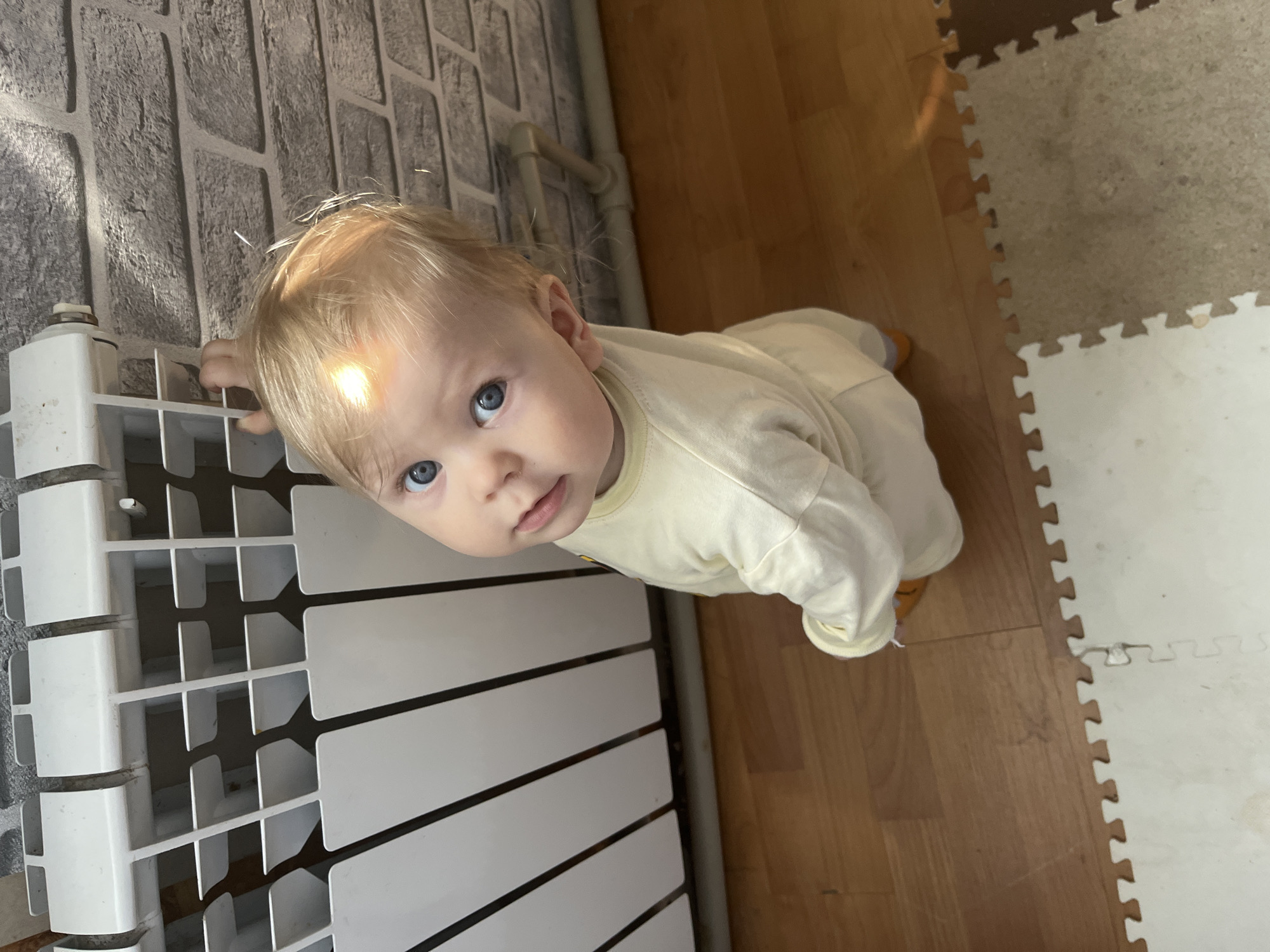 An overhead, slightly Dutch-angled shot captures a baby on all fours, looking directly upwards at the camera in an indoor setting in Vesyoloye, Russia. The baby, with fair blonde hair and striking blue eyes, wears a light yellow long-sleeved onesie or romper. A bright shaft of natural sunlight illuminates the top of the baby's head and the textured gray wall behind, creating a noticeable glare. The baby's right hand rests on the top of a white, modern-style heating radiator that occupies the lower left portion of the frame. The radiator is positioned against a gray wall resembling brickwork, with two light-colored pipes visible behind it. The floor is a mix of warm-toned wooden floorboards on the right and interlocking foam play mats in light gray/beige and white on the far right. The mats suggest a child-friendly area. The baby's expression is one of curiosity and direct engagement with the viewer. The strong natural light indicates it is daytime. There is no visible text.