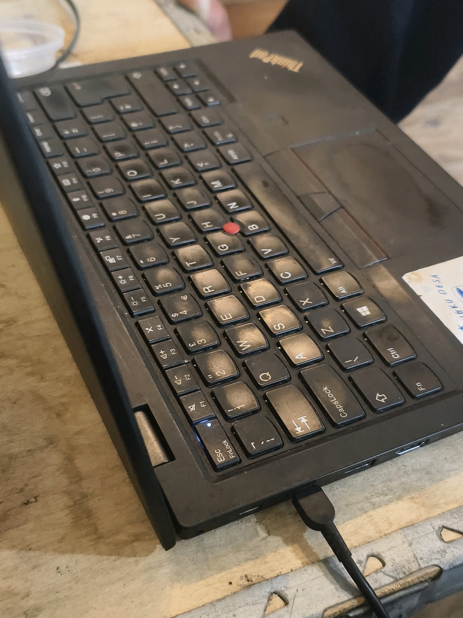 The image shows a close-up, angled view of a black Lenovo ThinkPad laptop keyboard with a USB-C cable plugged into its side. The laptop rests on a light-colored, textured surface, possibly a wooden table. The keyboard shows wear, particularly on the keys, indicating use. The "ThinkPad" logo is visible on the palm rest. A small, clear plastic container is partially visible in the upper left corner. A sticker with text, including "JIBU DESA," is attached to the laptop's chassis near the top right. The lighting suggests an indoor environment. The scene is devoid of any specific identifiable landmarks or details that would pinpoint its location to Muara Bulian, Indonesia.