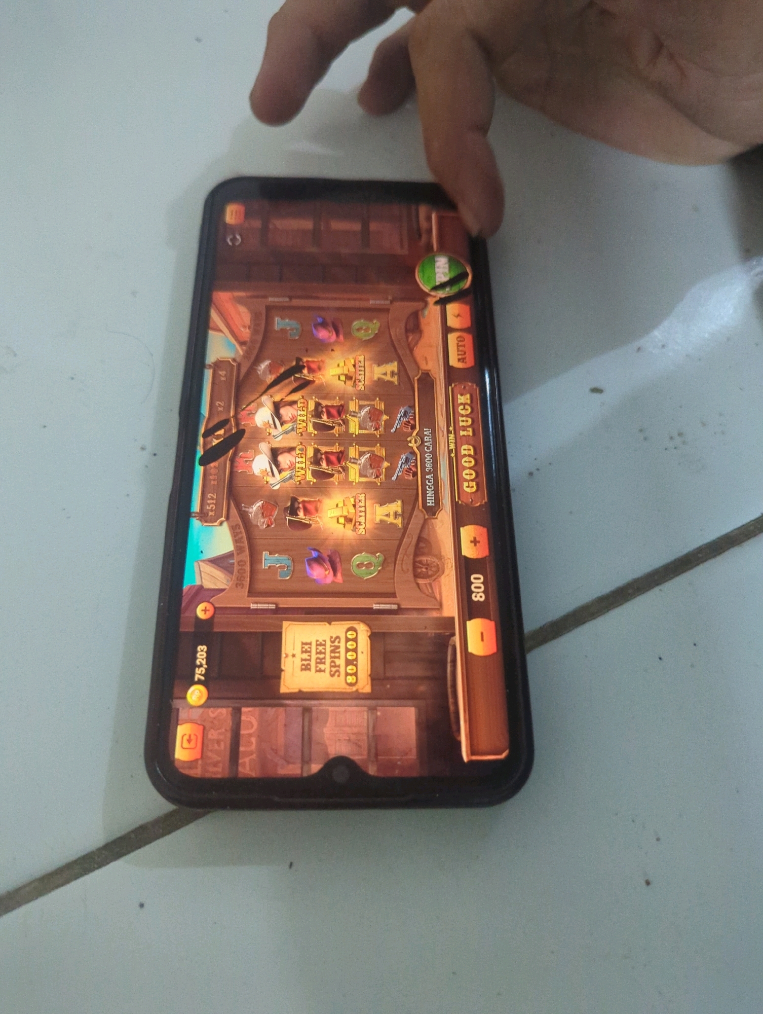 A hand is partially visible at the top of the image, touching the screen of a smartphone. The smartphone displays a slot machine game with a Western theme. The game features images of cowboys, revolvers, and card suits. There are buttons for "SPIN" and "AUTO," as well as options to adjust the bet. A text overlay on the game reads "GOOD LUCK" and "HINGGA 3600 CARA!". In the lower right corner of the screen, there's a notification for "BLEI FREE SPINS 80,000". The phone is lying on a tiled floor, which appears to be in an indoor setting. The lighting suggests it is daytime. The image does not provide any specific information about the city of Batu Belaman, Indonesia, or any other geographical location.