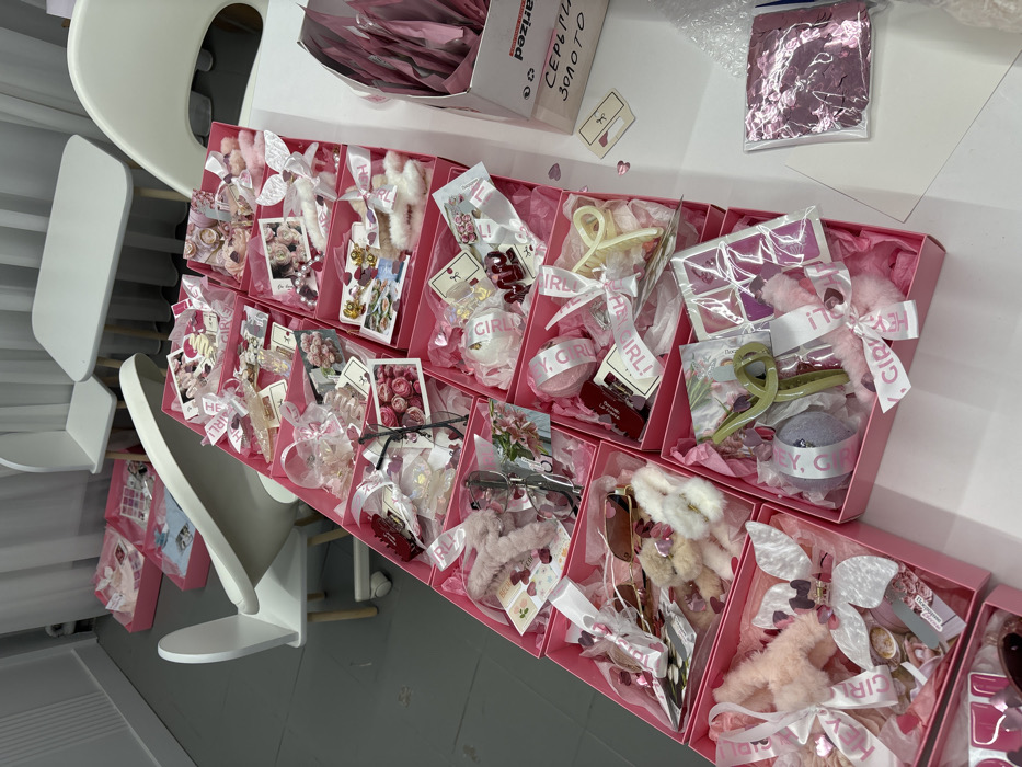 The scene depicts an indoor commercial or preparation area in Krasnodar, Russia. A white rectangular table in the foreground is largely occupied by multiple rows of open pink cardboard boxes. Each box contains an assortment of small fashion accessories and beauty products, predominantly in pink and light tones. Specific items include various hair clips (claw clips, barrettes), faux fur headbands, scrunchies, sunglasses, clear prescription glasses, sets of artificial nails, small makeup palettes, and cylindrical items resembling bath bombs. Many items are nestled in pink tissue paper, secured with white ribbons (some printed with "IT'S A GIRL!"), and accompanied by decorative cards featuring floral or heart motifs. Small pink heart-shaped confetti is scattered across the table surface and within some boxes. To the right, a partially visible cardboard box displays the Russian text "СЕРЬГИ ЗОЛОТО" (EARRINGS GOLD) and contains numerous individually wrapped pink items. The background reveals a light-colored room with light gray flooring, furnishing several white children's chairs with wooden legs and a small white rectangular table. White curtains or blinds are visible along the back wall. No individuals are present in the image.