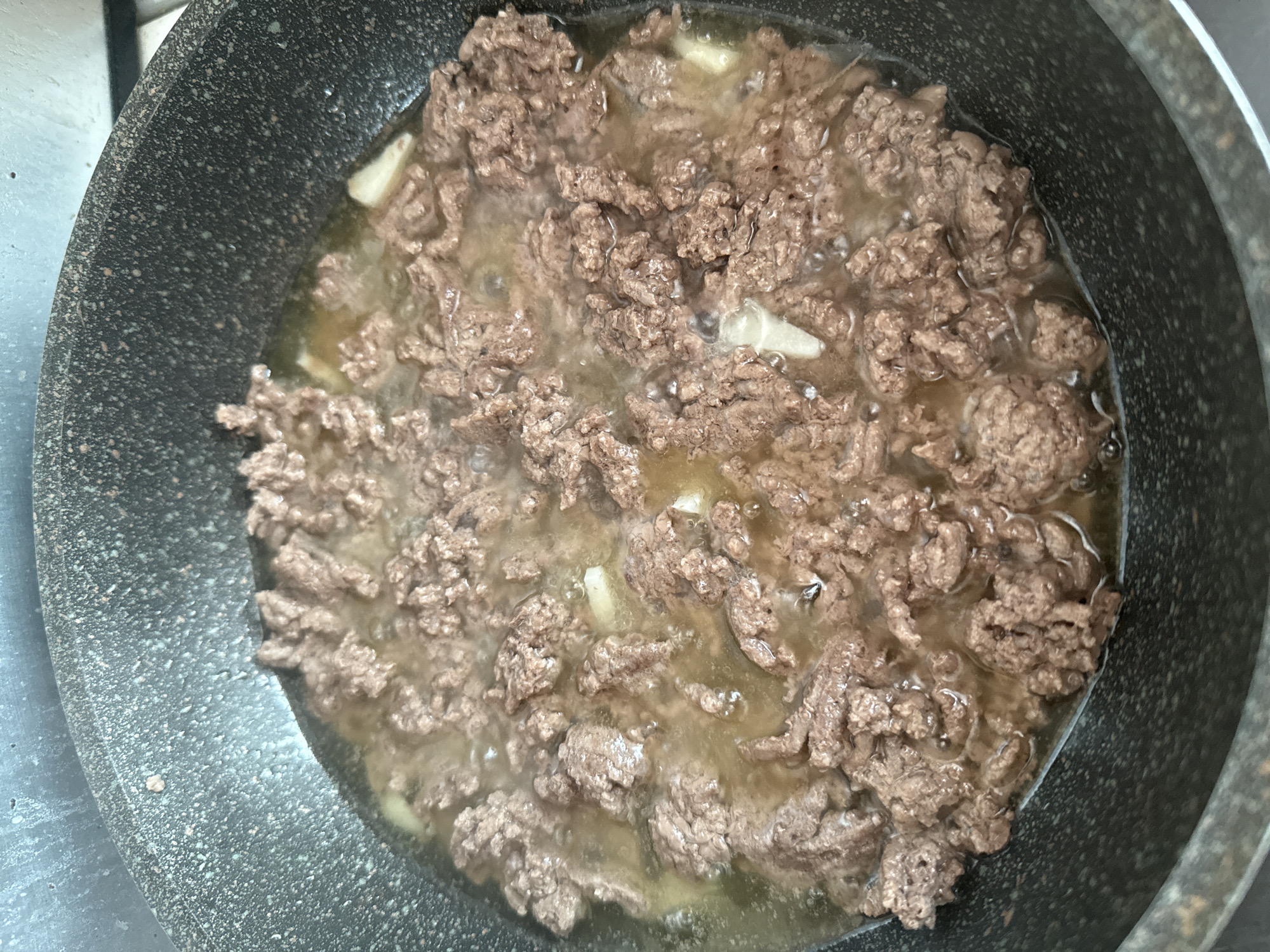 The image shows a close-up, overhead view of ground meat cooking in a speckled black non-stick pan. The meat is browned and appears to be simmering in its own juices and possibly some added liquid, creating a moist environment with visible steam rising. Small pieces of what appear to be garlic are interspersed with the meat. The pan has a slightly textured, granular surface, typical of many modern non-stick cookware. The setting is a kitchen, indicated by the presence of a stovetop surface visible in the upper left corner of the image. There are no people or specific time-of-day or weather cues. The overall impression is one of food preparation in progress.