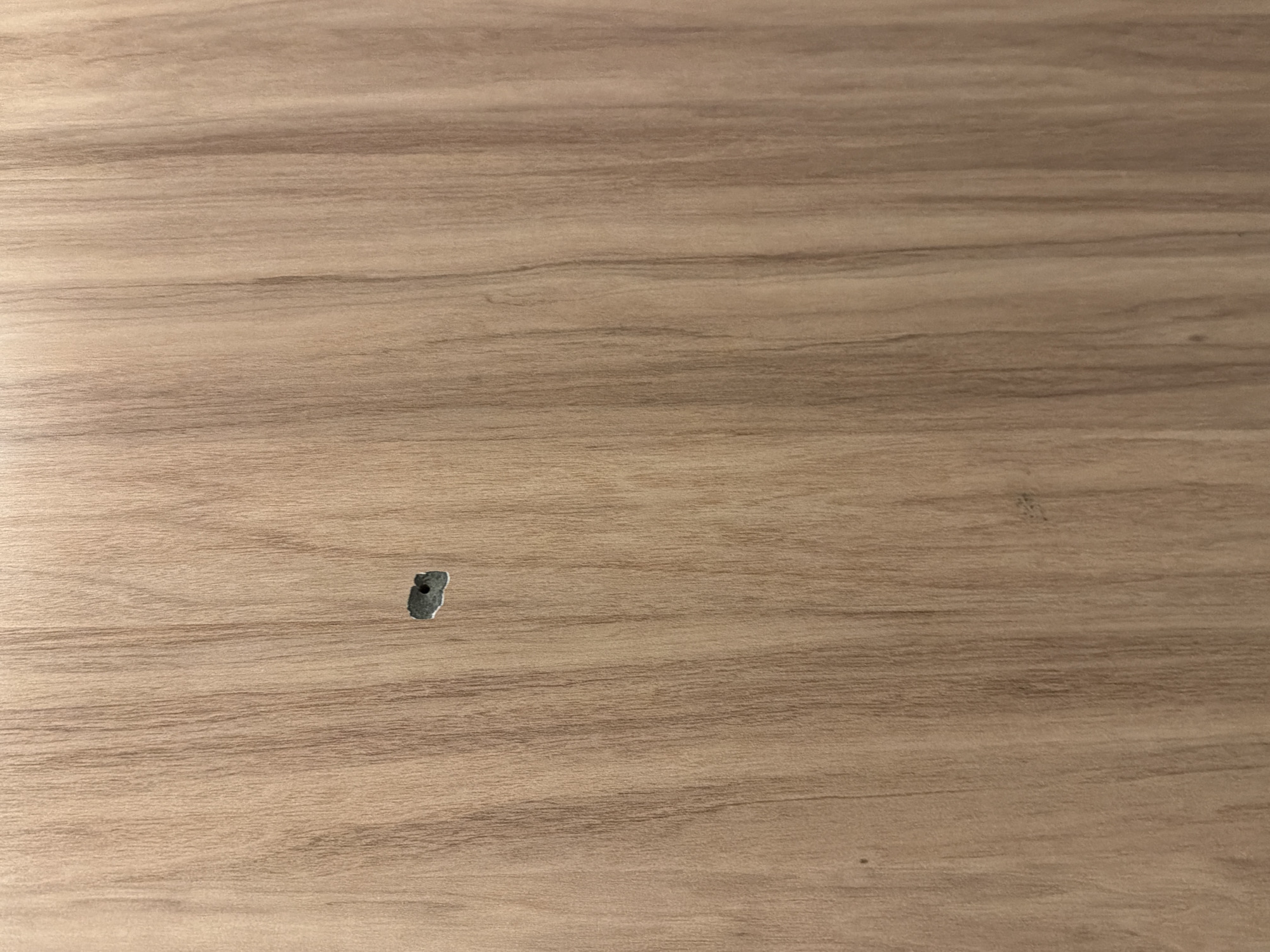 A close-up, eye-level shot shows a wooden surface with a small, irregular hole punched through it. The wood grain is predominantly vertical, with varying shades of light brown and beige. The hole, located near the center of the frame, is dark gray and appears to be a clean puncture. There are no other discernible objects or subjects in the image, and the background is uniformly the wooden surface. This image was taken in Sofia, Bulgaria.