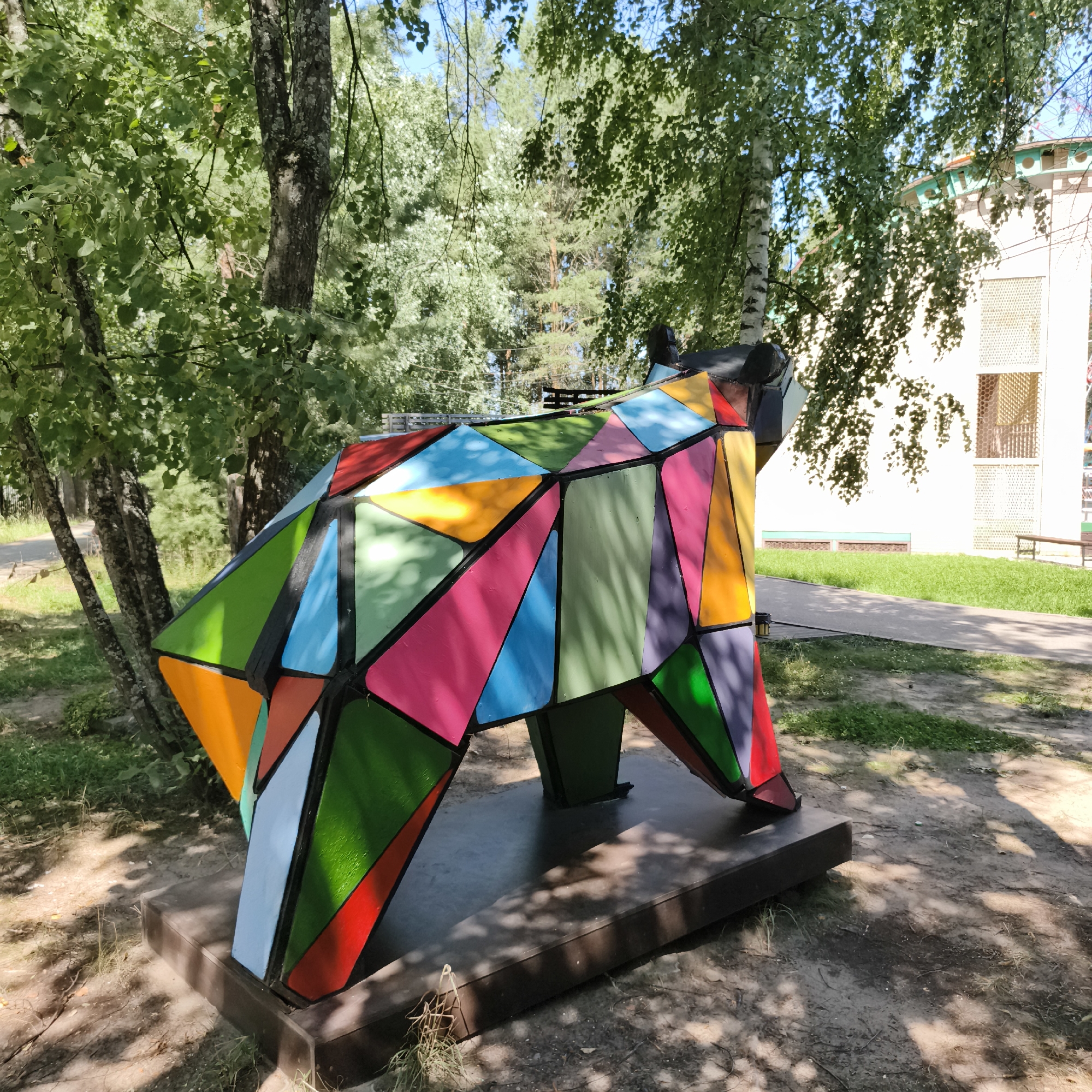 A large, abstract, multi-colored sculpture resembling a bear stands on a dark base in a park setting. The sculpture is composed of geometric shapes, predominantly triangles and quadrilaterals, painted in vibrant colors such as red, orange, yellow, green, blue, pink, and purple. The sculpture is positioned outdoors with trees and greenery surrounding it. In the background, a light-colored building with windows and a green trim is visible. The ground around the sculpture is covered in dirt and grass, with shadows from the trees dappling the scene. The image appears to have been taken during the daytime on a sunny day. The location is the Park of Culture and Leisure named after P. I. Chaikovsky in Chaikovsky, Russia, specifically near the area by the building of the House of Sports.