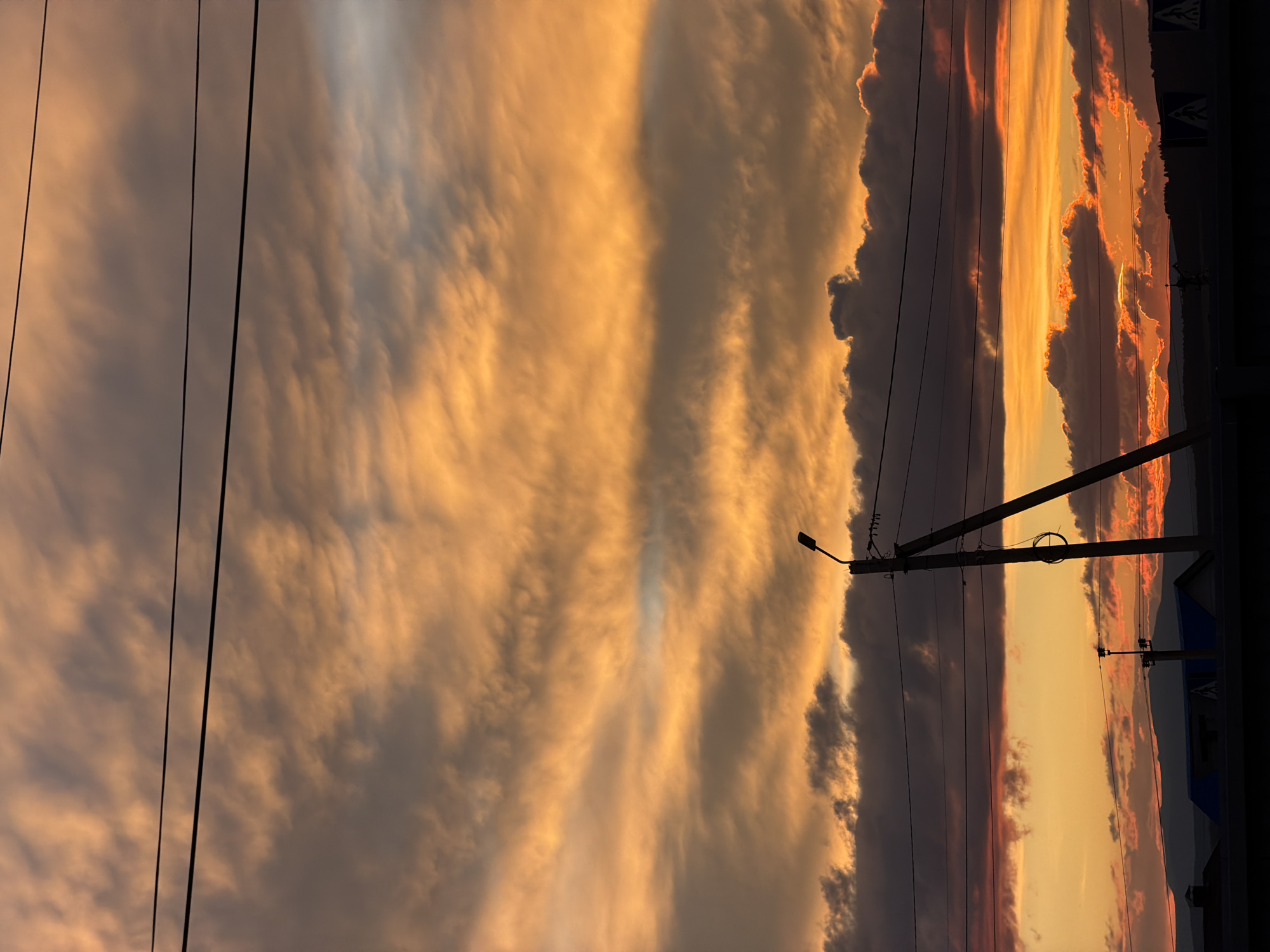 A vertical, outdoor, eye-level shot captures a dramatic sunset over a rural landscape. The sky is filled with vibrant orange and yellow clouds, tinged with grey and hints of blue, creating a striking contrast. Power lines stretch horizontally across the upper portion of the frame. In the middle ground, a tall power pole with a streetlight stands to the right, silhouetted against the colorful sky. Another smaller utility pole is visible further to the left. The lower part of the image shows a dark silhouette of buildings and trees along the horizon, with distant mountains partially visible. A faint suggestion of a road with pedestrian crossing signs is present at the bottom right. The overall mood is atmospheric and awe-inspiring.