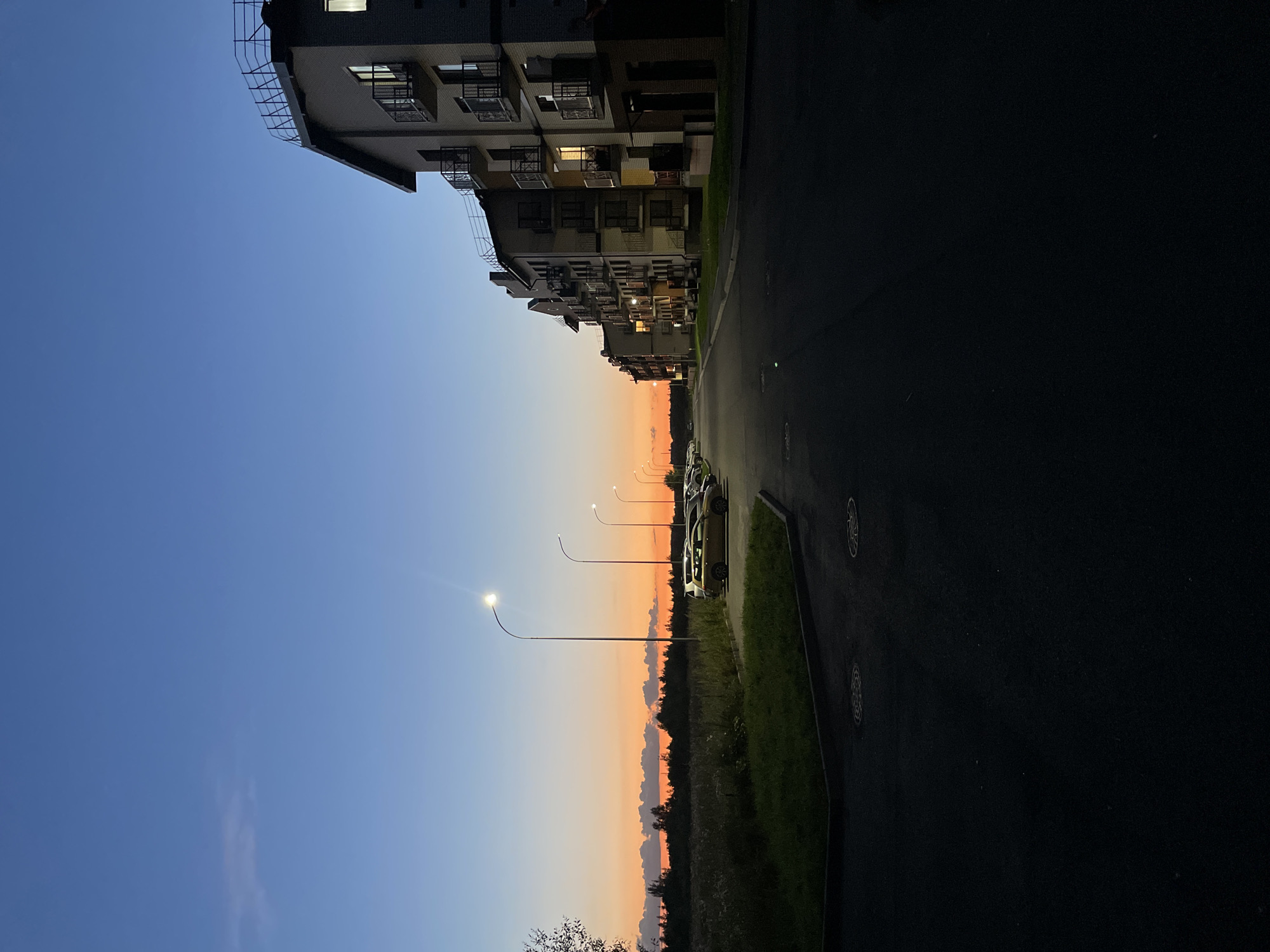 The image depicts a dusk scene in the settlement of Korenevo, Russia, featuring a row of modern apartment buildings on the right side of a paved road. The sky transitions from a deep blue at the top to a vibrant orange and yellow hue near the horizon, indicative of sunset. Several streetlights are illuminated along the road, casting pools of light. Parked cars are visible in a parking area on the left, adjacent to a grassy verge and a field with sparse vegetation and trees in the background. The architecture of the buildings suggests a residential complex. The scene is devoid of human presence.