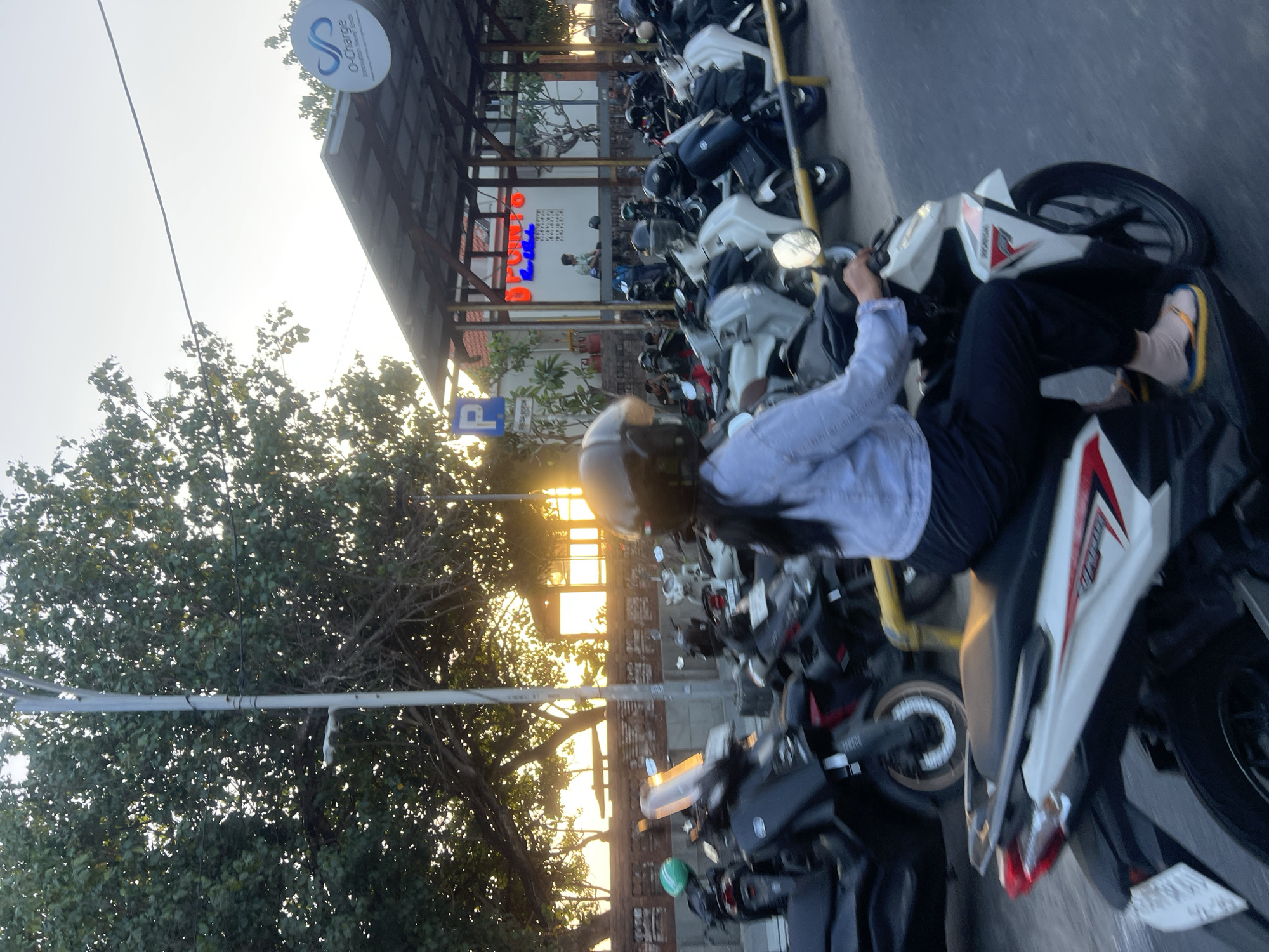 In Kuta, Indonesia, a person wearing a helmet and a light blue long-sleeved shirt rides a white motorcycle with red and black accents. They are wearing dark pants and yellow and blue flip-flops. The motorcycle has a license plate that is partially visible. The scene is a street, possibly a parking area, with numerous other motorcycles parked. In the background, there is a covered structure with a sign that says "O-Charge" and a symbol resembling infinity. There is also a blue parking sign with a "P" symbol. Trees with lush green leaves are visible on the left side of the image, and the sun is setting, casting a warm glow over the scene. The ground appears to be asphalt.