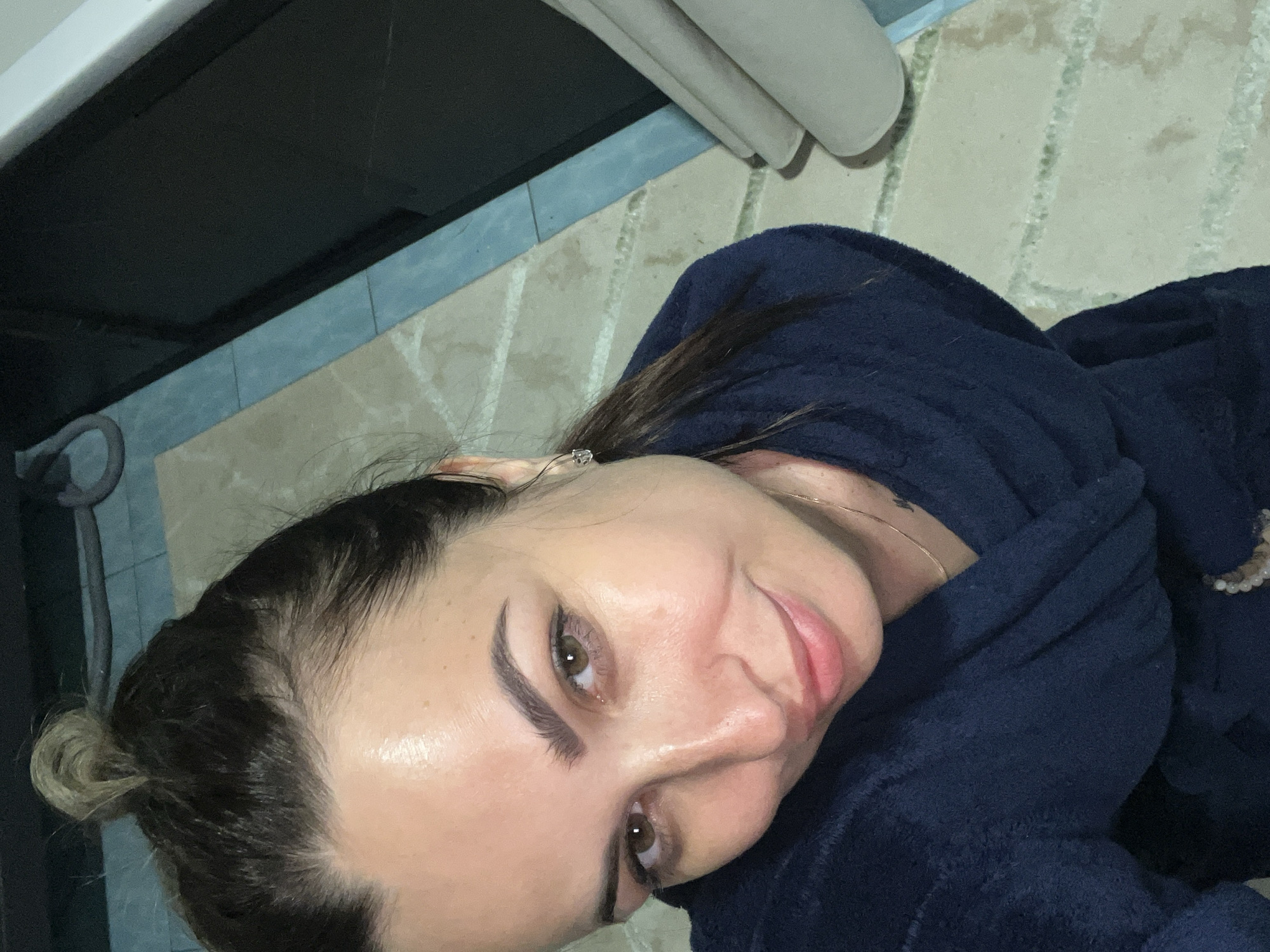 This is a close-up, low-angle selfie of a woman wearing a dark blue robe. She has her hair pulled back in a bun and is looking directly at the camera with a slight smile. Her makeup includes winged eyeliner and filled-in eyebrows. She is wearing a small diamond stud earring. The background appears to be a bathroom with tiled flooring and what looks like a dark countertop or cabinet. The lighting suggests it could be daytime, but there are no strong indicators of the time of day or weather. The overall emotion conveyed is relaxed and casual. There is no visible text in the image.