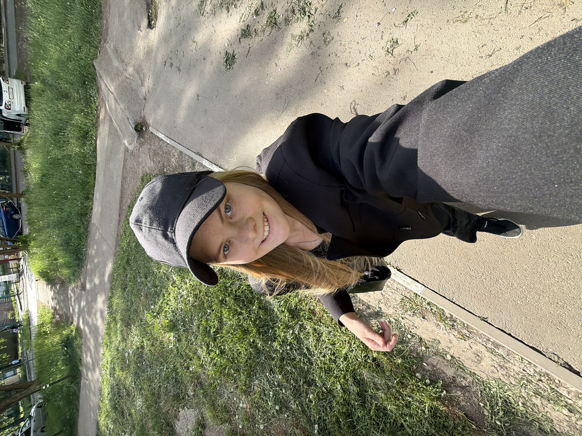 The image is a selfie of a young woman, likely taken in a park or green area within the city of Izhevsk, Russia. The woman is fair-skinned, with blonde hair partially obscured by a dark grey baseball cap. She is wearing a black blazer and dark trousers. She is smiling at the camera. The background consists of grass, trees, and a paved path. There are buildings and cars visible in the distance. The lighting indicates it is daytime.