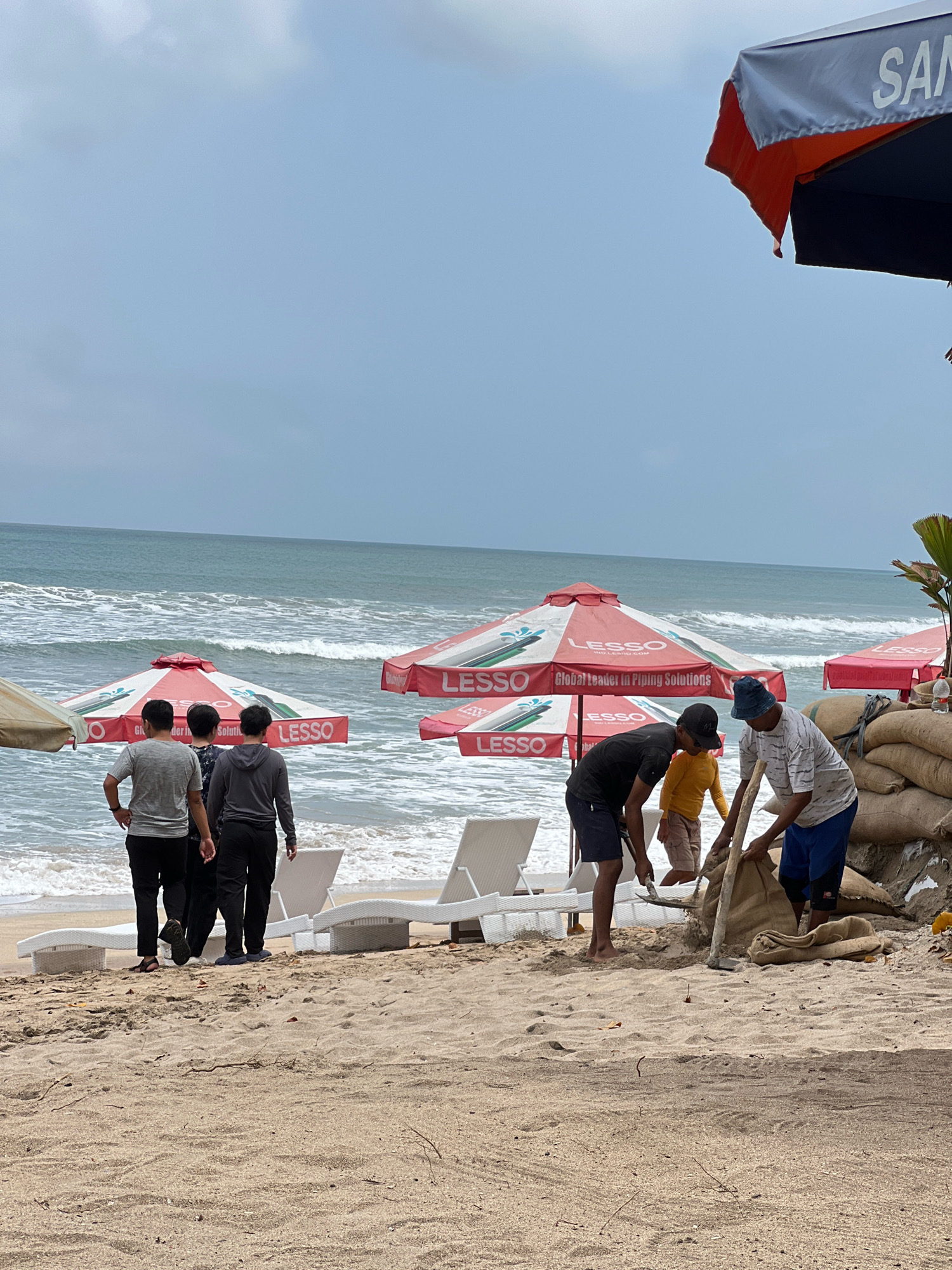A group of people are on a beach in Kuta, Indonesia, with the ocean in the background. In the foreground, three men are working with sandbags. One man is shoveling sand into a bag, another is holding a shovel and appears to be helping, and a third man is standing nearby, also appearing to assist with the sandbags. There are several large, brown sandbags stacked on the right side of the image. Several large, red and white beach umbrellas with the logo "LESSO" are visible on the beach, as are a few white lounge chairs. The ocean has moderate waves and the sky is overcast. The beach is sandy.