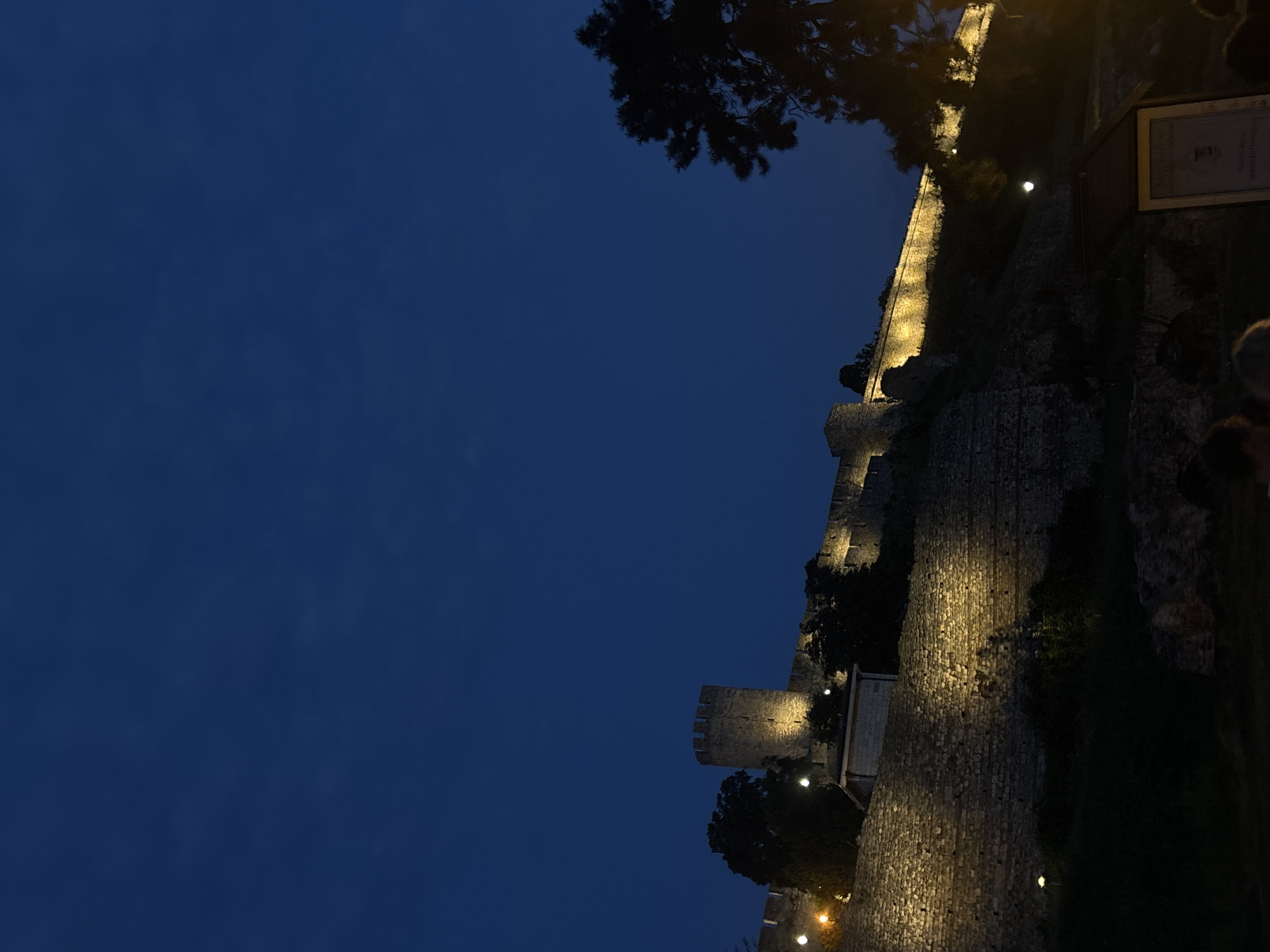 The image depicts a nighttime view of the Belgrade Fortress, locally known as Kalemegdan, in Belgrade, Serbia. The fortress walls, constructed from rough-hewn stones, are illuminated by warm spotlights, highlighting their texture and height. A prominent tower, part of the fortress's defensive structures, stands on the left side of the image, also lit by a spotlight. The sky above is a deep, dark blue, suggesting twilight or early night. Lush green trees are visible, some silhouetted against the illuminated walls and sky. In the foreground, the lower part of the image shows darker, less distinct areas, with hints of people's heads and shoulders visible, suggesting they are gathered at the base of the fortress. A wooden information stand with some text is also partially visible on the right. The scene conveys a sense of historical grandeur and atmospheric evening ambiance.