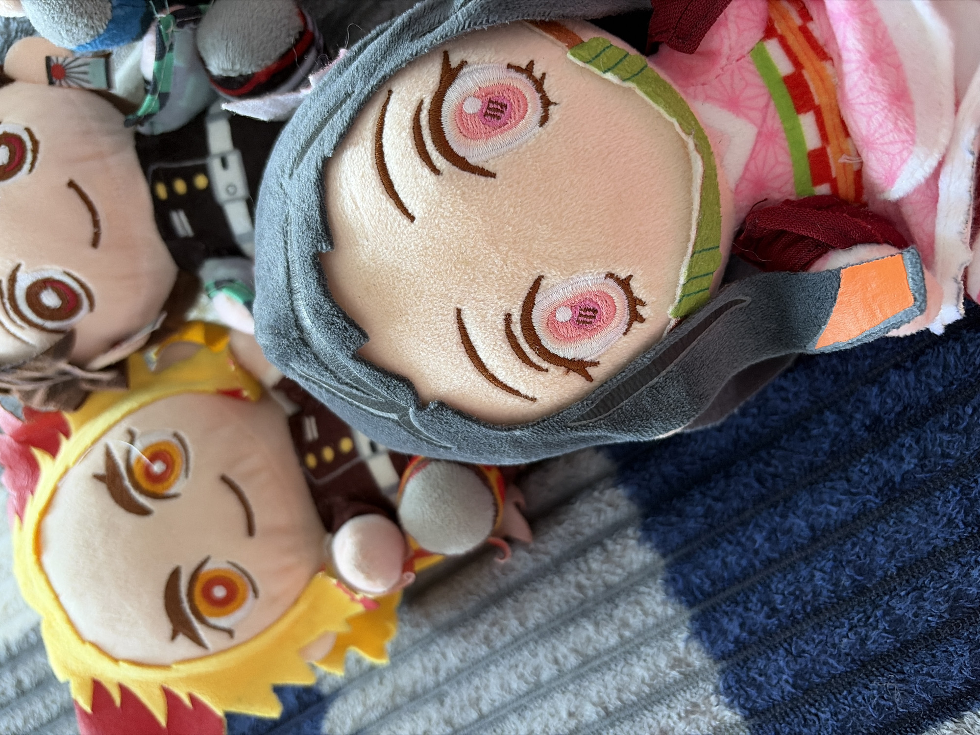 The image shows a close-up of several plush toys on a blue and gray striped fabric surface. The toys include Nezuko Kamado from Demon Slayer, with a gray hood and pink kimono. There is also a yellow-haired character, possibly Renkogu Kyoujuro, and another plush toy with white skin and brown eyes. The image is taken at a close range, with a shallow depth of field, making the background slightly blurred. No specific location in Chaikovsky, Russia, can be determined from the visual data.