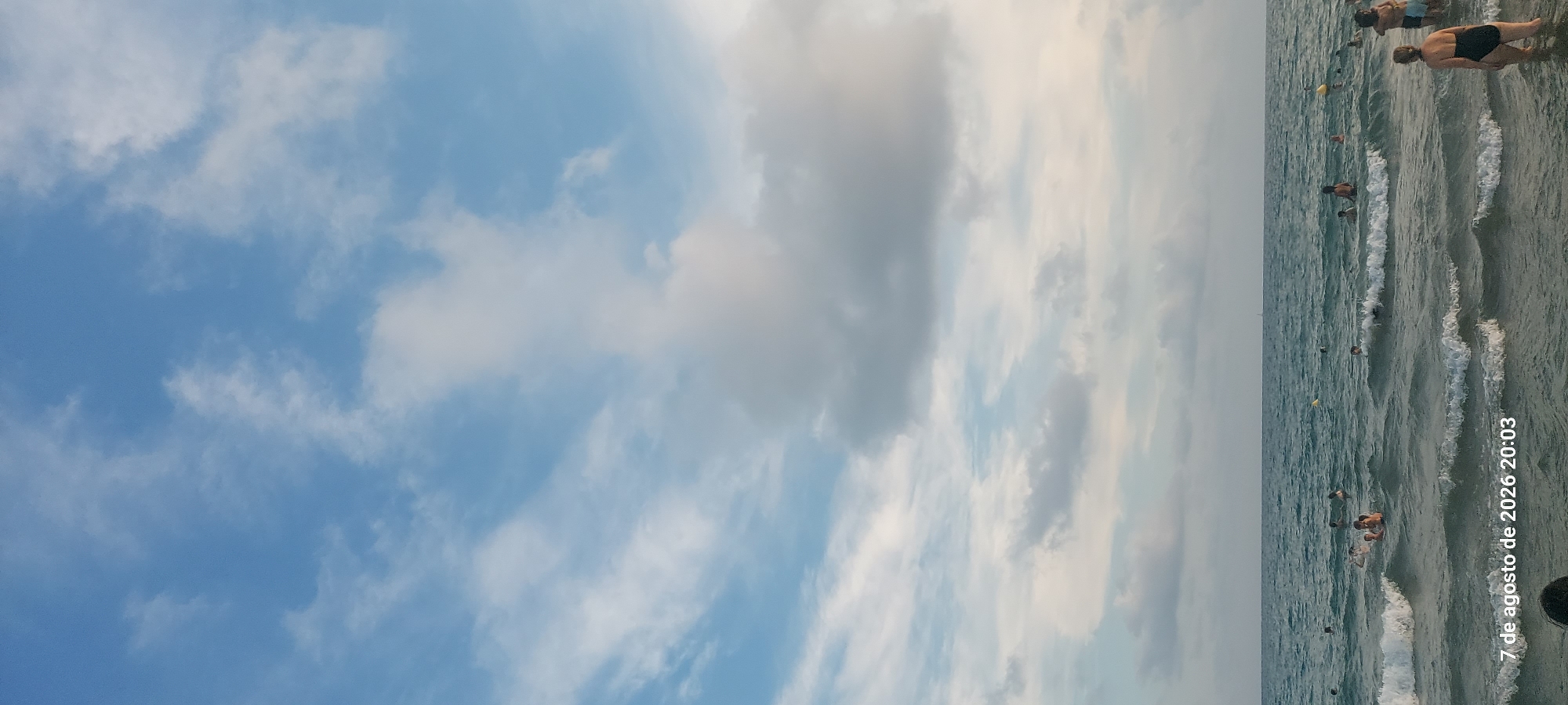 The image depicts a beach scene in Castelldefels, Spain, as indicated by the timestamp "7 de agosto de 2026 20:03". The vast sky above is filled with scattered white and gray clouds against a backdrop of blue. Below the sky, the Mediterranean Sea is visible, with moderate waves breaking near the shore. Several people are in the water, swimming and wading. Some are partially submerged, while others are further out. There are at least five visible individuals, with more implied by their presence in the water. A few yellow buoys are scattered in the sea. The lighting suggests it is late afternoon or early evening. In the foreground, the upper part of a person's head and shoulders, covered in dark hair, is visible at the bottom of the frame.