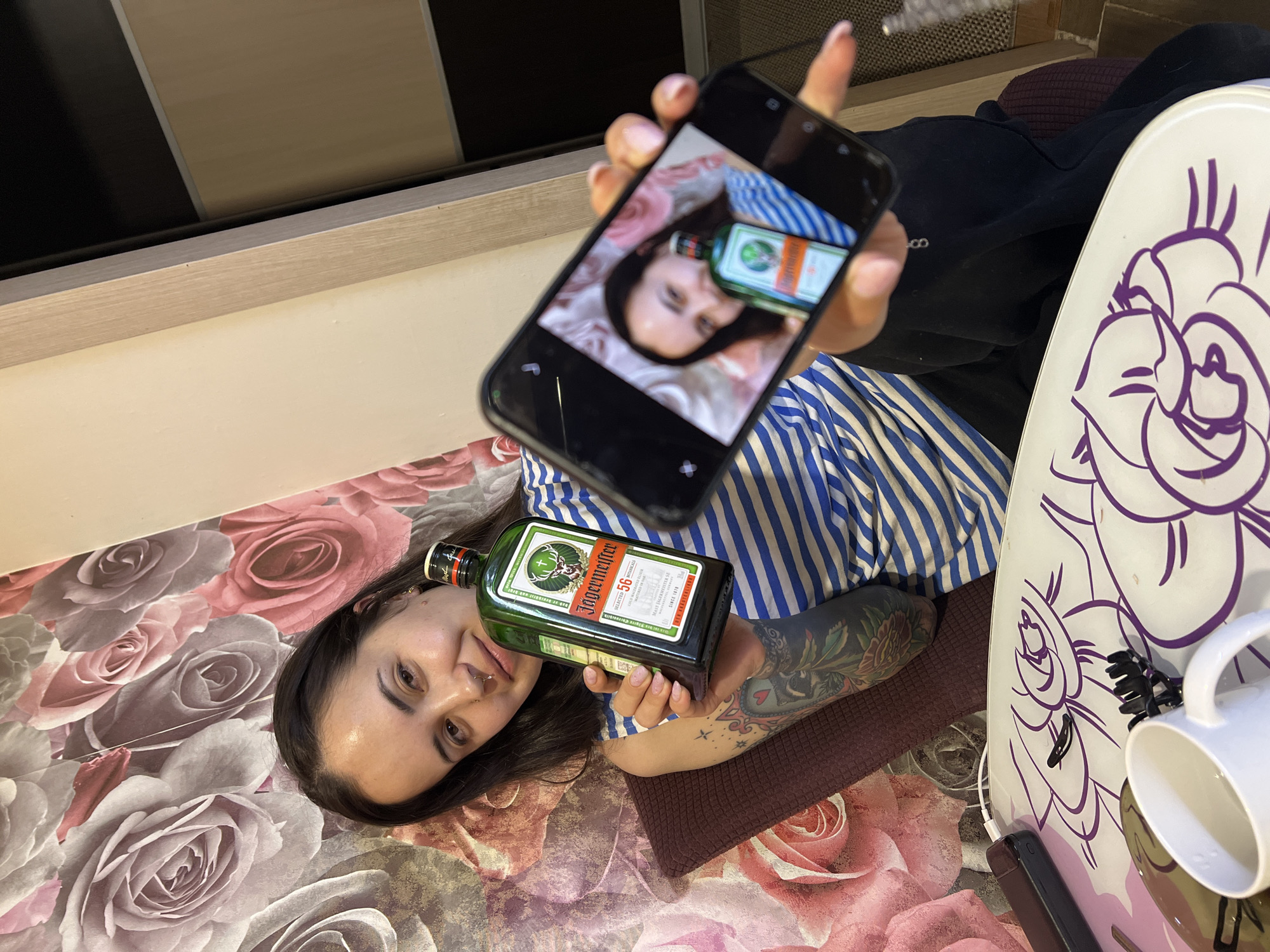 A young woman is positioned on a floral-patterned surface, potentially a bed or rug, holding a bottle of Jägermeister liqueur in her left hand. She is wearing a blue and white striped shirt. Her right hand holds a smartphone displaying an image mirroring the scene, featuring herself and the Jägermeister bottle. The background includes a light-colored wall or shelf. To the right of the woman is a white tray decorated with purple floral designs, and a white mug is on the tray. Additionally, there is a charging cable near the tray. The woman has dark hair, multiple piercings on her left ear, and a visible tattoo on her left forearm.