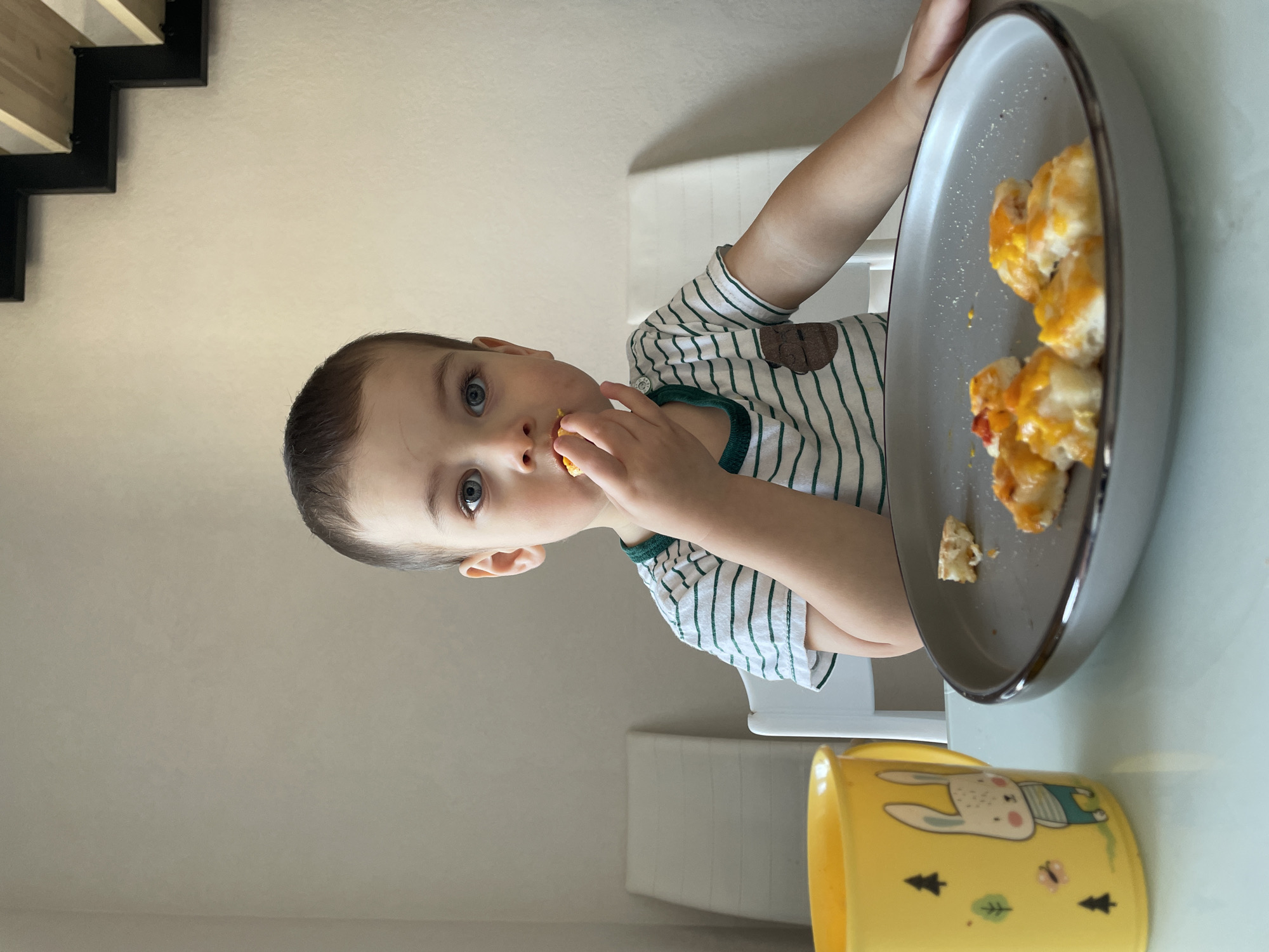 A young child with fair skin, short brown hair, and blue eyes is seated indoors at a light-colored table, consuming food. The child wears a white t-shirt with thin green horizontal stripes and a brown emblem on the left chest, looking directly into the camera while holding a small, orange-topped food item to their mouth with their right hand. On the table to the child's left, a yellow plastic mug featuring a cartoon rabbit, trees, and a butterfly stands next to a dark grey plate holding several small, irregularly shaped food pieces topped with melted orange-yellow cheese and red fragments. In the background, two white chairs with slatted backs are visible against a plain, light-colored wall. A modern staircase with dark metal risers and light wooden treads is partially visible in the upper right quadrant of the scene. The specific city of Izhevsk, Russia, cannot be determined from the visual evidence provided in the image.