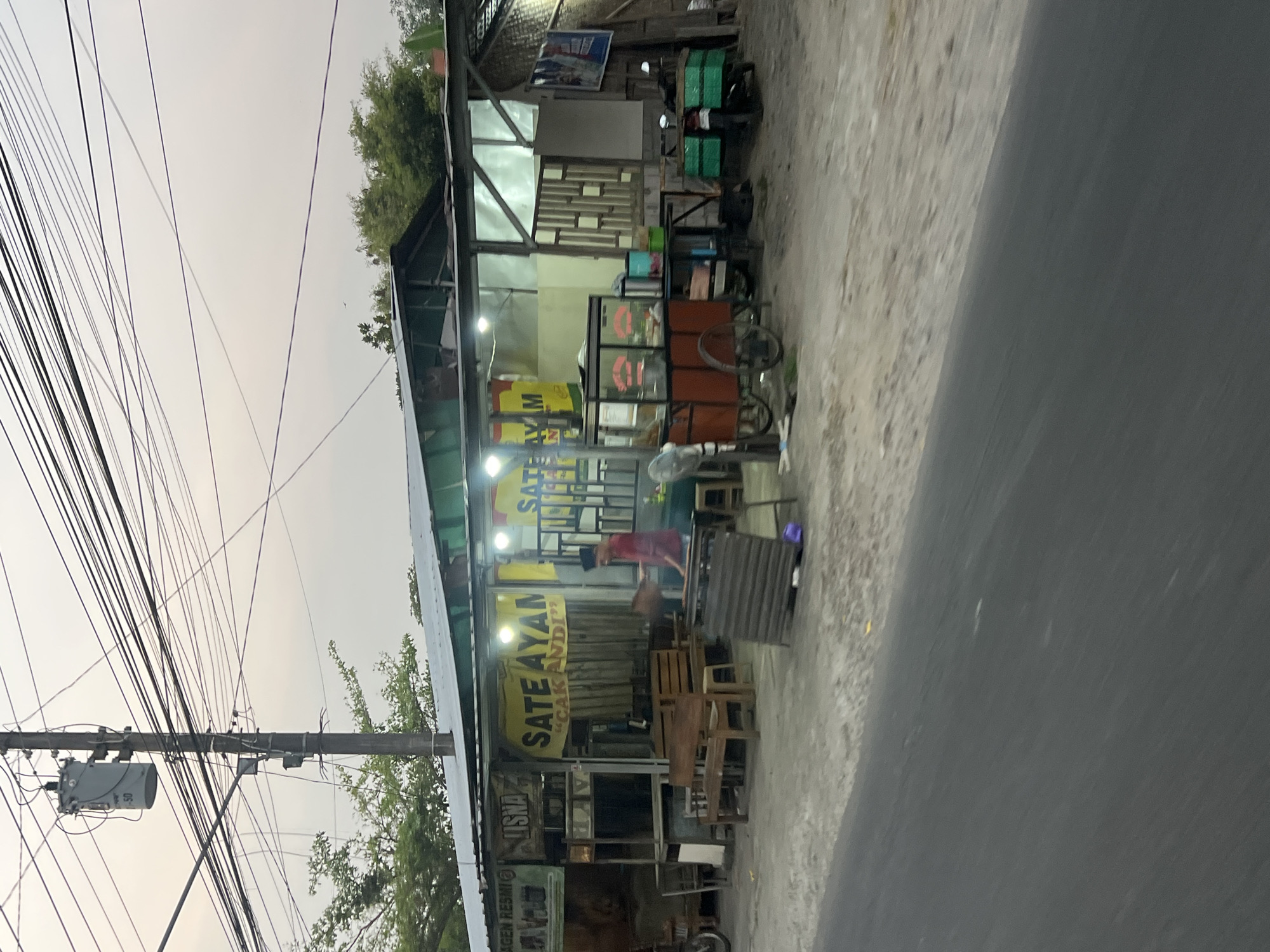 The image shows a street-level view of a small food stall or restaurant, likely in Sendangadi, Indonesia, based on the signage which includes "SATE AYAM" (chicken satay) in Indonesian. The establishment has a corrugated metal roof extending over a sidewalk area. A prominent yellow banner with "SATE AYAM" and "CAK ANDI" is displayed. In front of the building are several wooden benches and tables. To the right, there is a food cart with an orange frame and glass display, a metal fan, and a table with various containers. Further to the right, a motorcycle is parked with green boxes attached to the rear. A person wearing a red shirt and a black cap is visible near the food cart, appearing to be engaged in an activity related to the food preparation or service. Power lines crisscross the sky above, supported by a utility pole with a transformer. The ground in front of the establishment is a mix of dirt and asphalt. The overall scene depicts a typical roadside eatery in Indonesia.