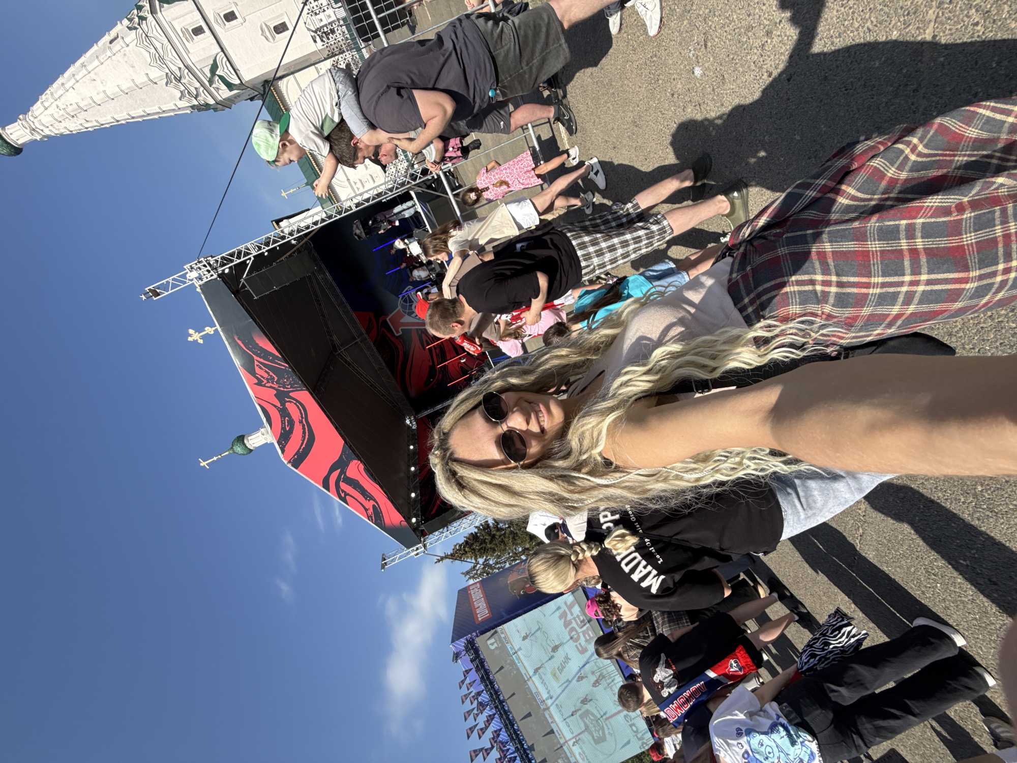 The image is a selfie taken outdoors in Заволжье, Russia. A blonde woman wearing sunglasses and a black sweatshirt is in the foreground. Behind her, there is a stage with red and black decorations. A crowd is gathered in front of the stage. To the left of the stage, a building with a spire is visible against a blue sky.