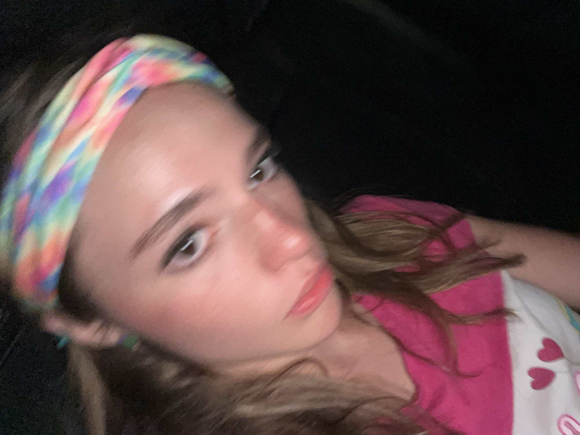 A young person with light skin is wearing a tie-dye headband and a pink and white striped shirt with hearts on it. They appear to be indoors, possibly in a car, as suggested by the dark background. The person is looking upwards and slightly to the right. The location is unknown, there is no evidence that the image was taken in San Bernardino Tlaxcalancingo, Mexico.