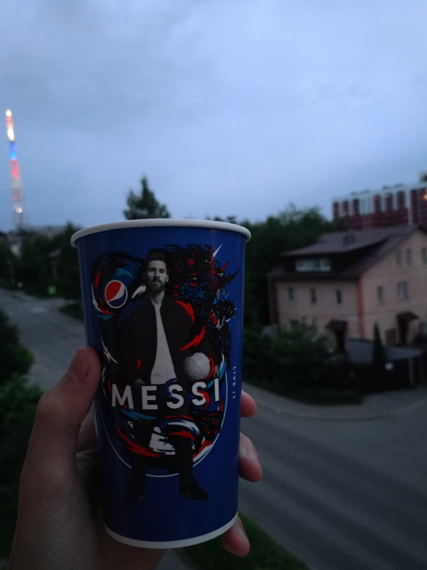 A person is holding a blue Pepsi cup with an image of Lionel Messi and a dragon. The cup is adorned with the Pepsi logo and the word "MESSI." In the background, a city skyline is visible with buildings and trees. A tall tower with colorful lights is also present in the distance. The sky is cloudy, suggesting either dawn or dusk. The scene appears to be an outdoor setting, possibly a balcony or rooftop overlooking a street. The specific location is the city of Petrozavodsk, Russia.