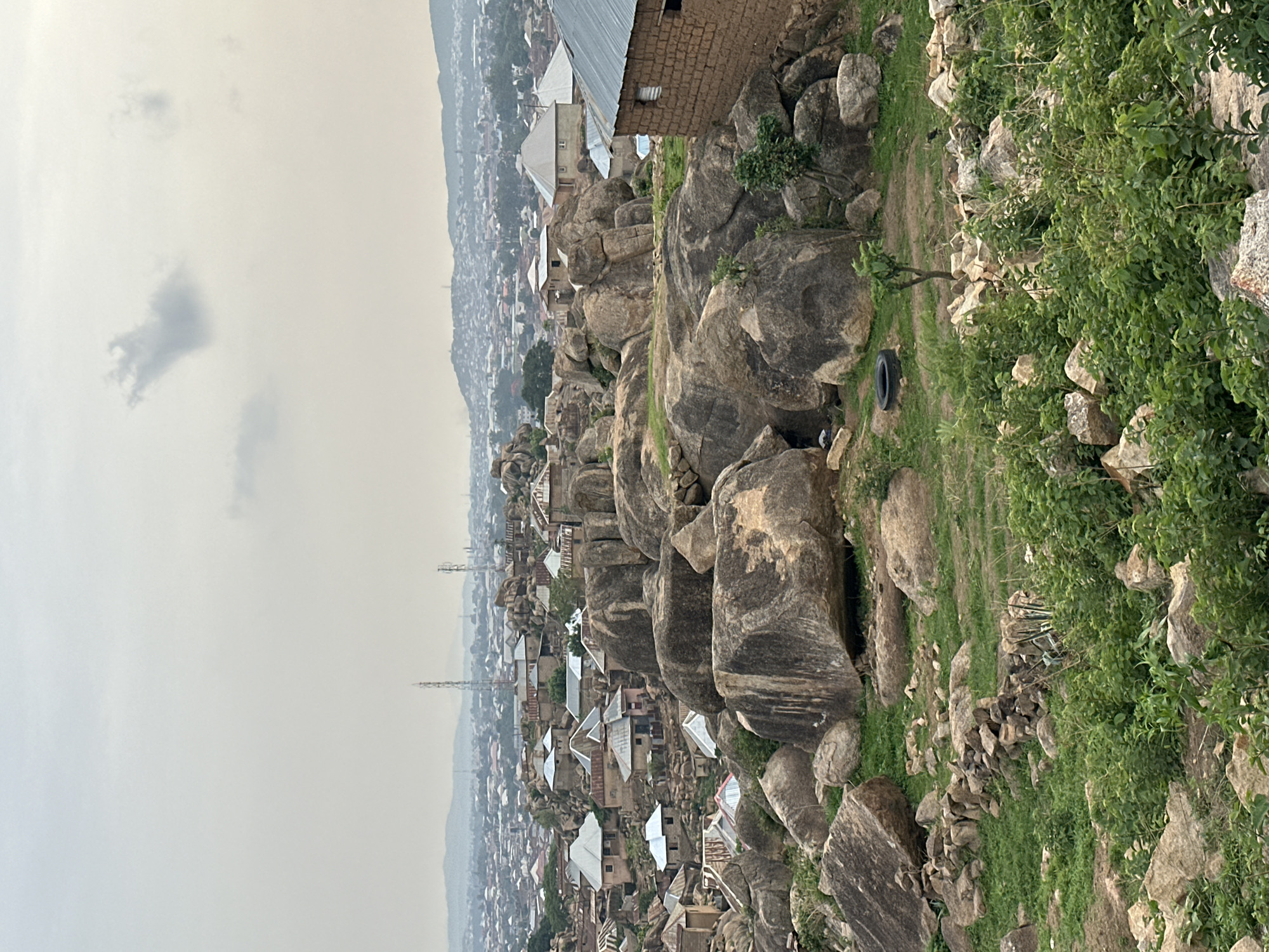 The image presents a panoramic landscape depicting a settlement integrated into a rugged, rocky terrain, characteristic of Jos, Nigeria. The scene is viewed from an elevated position, looking down upon the area.
In the foreground, large, weathered grey boulders and rock formations are prominent, interspersed with patches of green grass and dense bushes. A single black tire is visible resting on the ground among the rocks and sparse vegetation.
The midground reveals a village or residential area where houses are constructed directly among and on top of these natural rock formations. The buildings largely feature corrugated tin roofs, with some structures, such as a mud-brick house visible in the upper part of the foreground, showing more traditional construction methods. The architecture demonstrates adaptation to the challenging topography.
Further into the background, the settlement expands into a sprawling town or city, with numerous buildings stretching towards the distant horizon. Faint outlines of hills or mountains are visible in the far background. The sky is overcast and hazy with a few subtle clouds, suggesting diffused light, possibly indicating an early morning, late afternoon, or simply a cloudy day. Several tall, slender utility poles or communication towers rise above the distant urban landscape. No individuals or specific activities are discernible in the scene. There is no visible text.