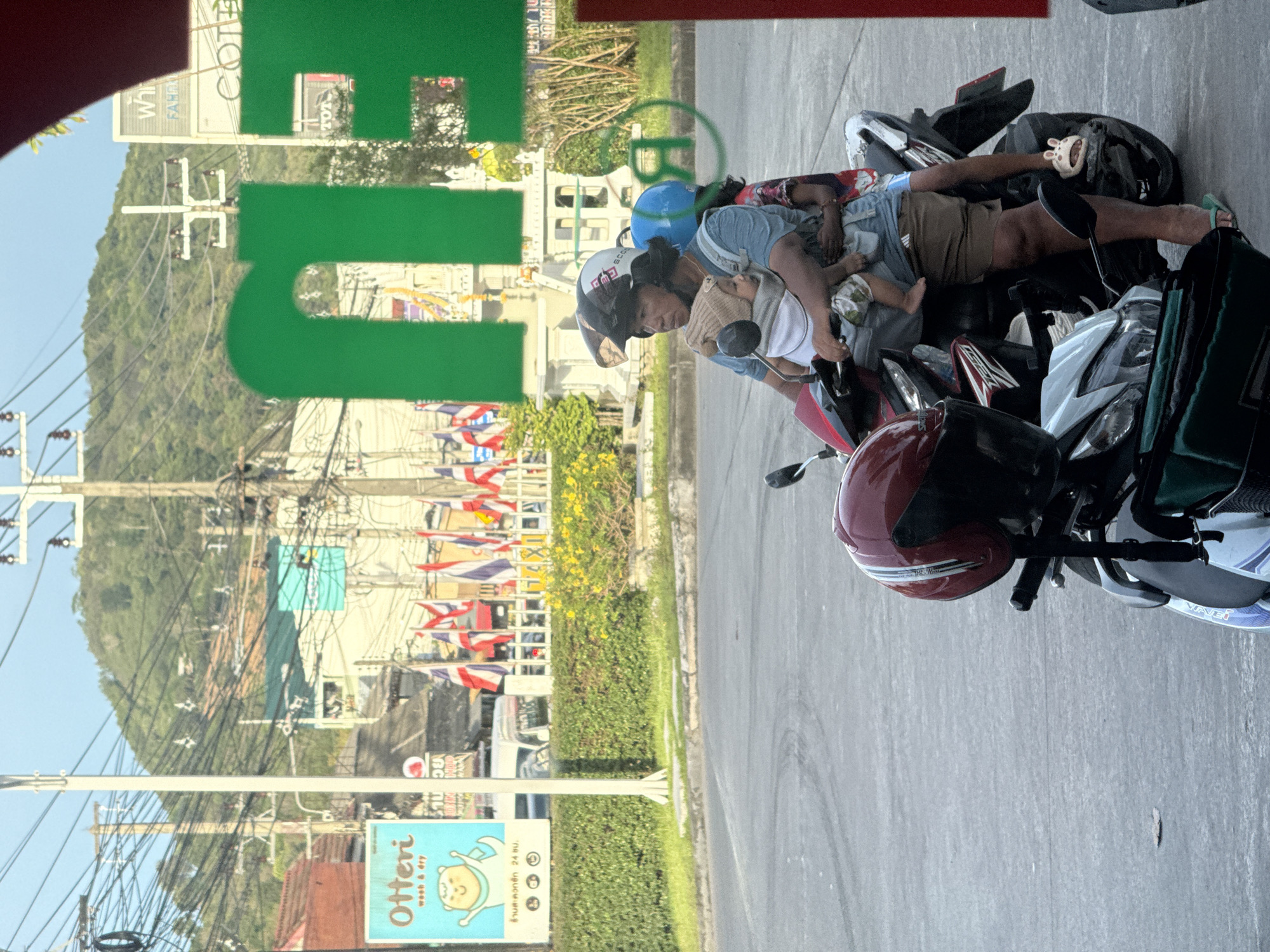 In Baan Bo Phut, Thailand, a man, woman, and two children are on a motorcycle. The man is wearing a helmet and carrying a baby in a front carrier. The woman is behind him, also wearing a helmet, and holding another child. The motorcycle is parked on a paved surface, possibly a street or parking lot. In the background, there are buildings, trees, and a hill. There are also several Thai flags visible. The overall scene suggests a typical day in a bustling Thai town.