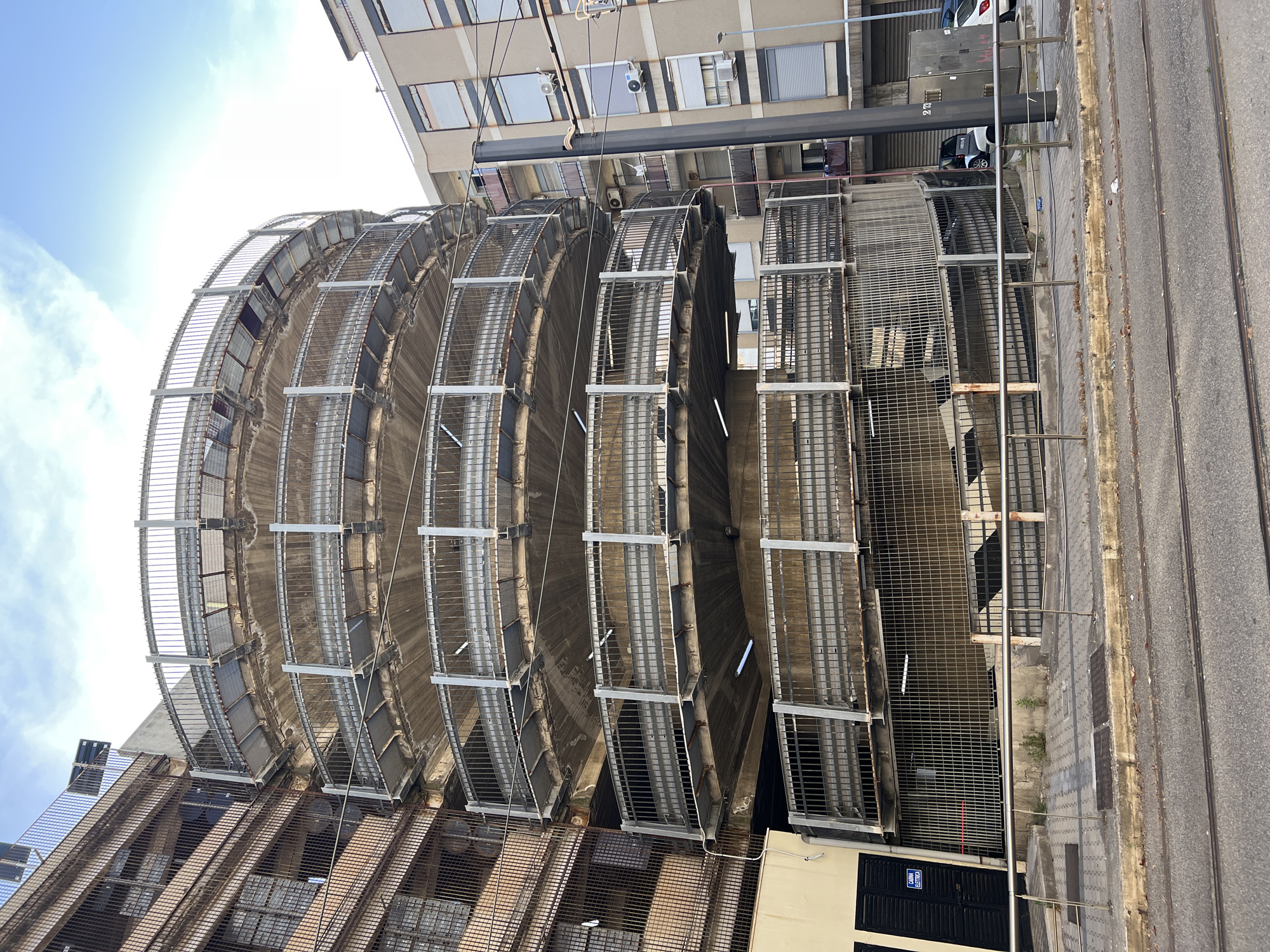 This image depicts an urban street scene in Messina, Italy, specifically featuring the **Parcheggio Cavallotti (Cavallotti Parking Garage)** near the intersection of Via Tommaso Cannizzaro and Viale Europa. The primary subject is a large, circular, multi-story spiral ramp structure, likely a parking garage, constructed from weathered concrete and metal. This structure occupies the center and left-center of the frame. It displays approximately six visible levels, each composed of a curved concrete deck and an exterior protective barrier system of horizontal metal railings and vertical metal mesh or grates. The concrete shows signs of age and discoloration, while the metal components exhibit rust and weathering. Fluorescent tube light fixtures are visible attached to the underside of several concrete decks. On a lower level, partially visible through the grate, a small, two-door vehicle (possibly a Smart car) is parked within the garage. The lowest visible level features a large access opening secured by vertical metal bars. To the left of the spiral structure stands an older, multi-story residential building with a facade of light brown brick or exposed concrete. Its numerous rectangular windows are protected by rusty, horizontal metal grates. The ground floor portion of this building, closer to the viewer, is painted a light yellow/cream color and features dark brown or black closed shutters. A blue rectangular sign with white text reading "CABINA ELETTRICA" (Electrical Cabin) is affixed to the yellow wall. Air conditioning units are visible on its exterior. To the right of the spiral structure, a more modern multi-story apartment building is visible, featuring a light beige/brown stucco facade. This building has multiple balconies with dark railings, some containing visible objects or draped textiles. Many windows are fitted with closed external roller shutters, and external air conditioning units are mounted below various windows. A tall, thick, black utility pole stands prominently in front of this building, partially obscuring it, with multiple electrical wires extending horizontally from its upper portion. In the immediate foreground, two parallel metal tram tracks are embedded in a worn, paved surface, running horizontally across the frame. Behind the tracks, a concrete sidewalk with a checkered pattern is visible, appearing somewhat discolored and uneven, with small patches of weeds growing through cracks. A low, silver-colored metal railing delineates the edge of the sidewalk. On the right side of the sidewalk, near the black utility pole, two dark gray utility boxes are present. Scattered minor debris, including