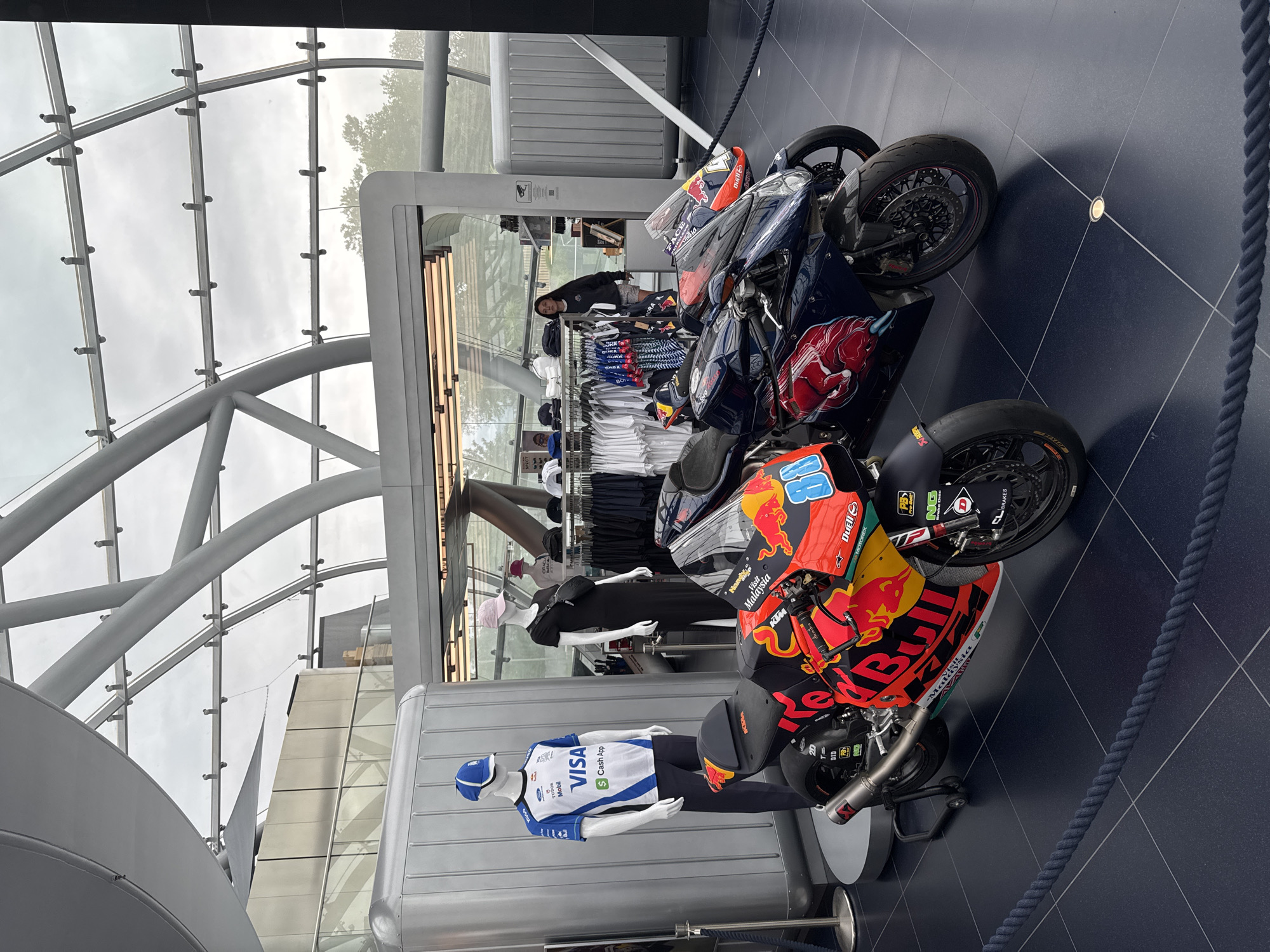 The image depicts the interior of a modern building, likely a retail space or exhibition area, with a curvilinear glass roof and metal supports. In the foreground, two racing motorcycles are displayed on a dark, tiled floor. The motorcycle on the left is predominantly orange and red with the Red Bull logo prominently displayed. The motorcycle on the right is dark blue with red accents, featuring a stylized bull graphic.
To the left of the motorcycles, there are mannequins dressed in sportswear. One male mannequin wears a blue and white racing jersey with "VISA" and "Cash App" logos, along with a blue cap. Adjacent to it, a female mannequin is dressed in a long black dress and a crossbody bag, wearing a pink cap.
Behind the mannequins and motorcycles, a retail display area is visible, with racks of clothing, hats, and merchandise. A young woman with dark hair and wearing a grey sweatshirt and shorts is browsing the merchandise. The overall environment suggests a promotional event or a dedicated retail space for a racing team or brand, possibly related to motorsports given the presence of the racing motorcycles. The building's architecture suggests a contemporary design, and the mention of Salzburg, Austria, places the scene in a specific European city.