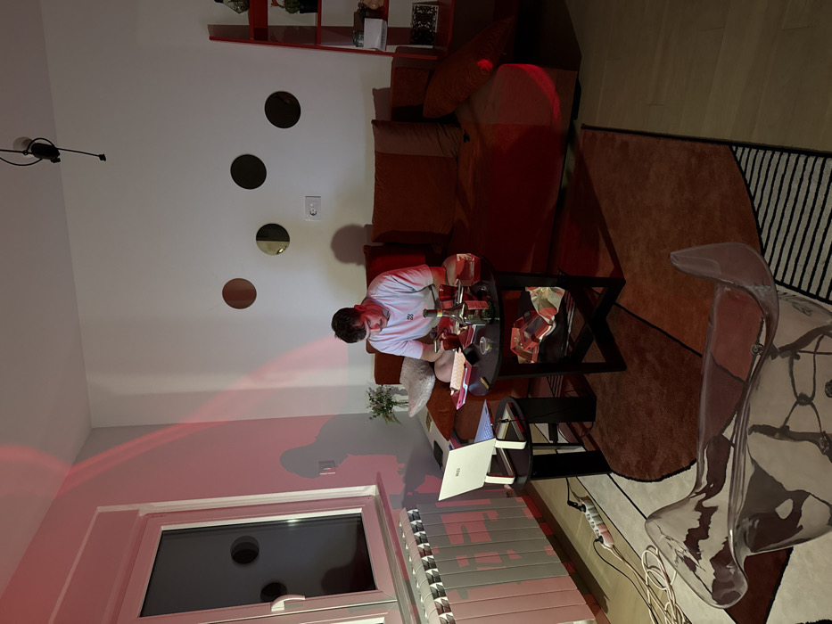 A person sits at a dark coffee table with numerous objects, possibly bottles, on it. A laptop is on a lower shelf of the table. A brown couch is behind the person, and four circular objects are on the wall. Red light is prominent in the lower left of the frame, near a window with blinds. A clear, ornate chair is on a rug with black and white stripes.