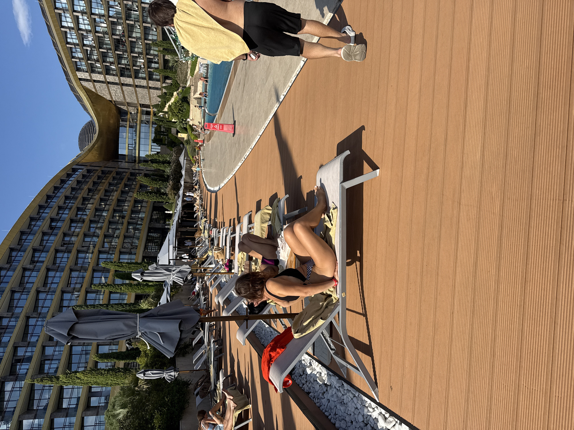 This image depicts a sunny day at a resort in Simeiz, Ukraine, likely an outdoor area adjacent to a swimming pool and a large, modern hotel. In the foreground, a woman in a black and white striped bikini relaxes on a lounge chair on a wooden deck. A red garment is draped over the armrest of her chair. To her right, another woman in a purple bikini is also lying on a lounge chair. Further in the background, several other lounge chairs are visible, some occupied by people.
A man wearing black shorts and a yellow towel draped over his shoulder is walking away from the camera on the wooden deck. In the middle ground, a swimming pool with clear blue water is visible, with people swimming and relaxing around its edge. The resort features a distinctive, curved, modern hotel building with many windows and balconies. Cypress trees are scattered throughout the grounds, adding greenery to the scene. The overall impression is one of a luxurious and relaxing vacation setting.