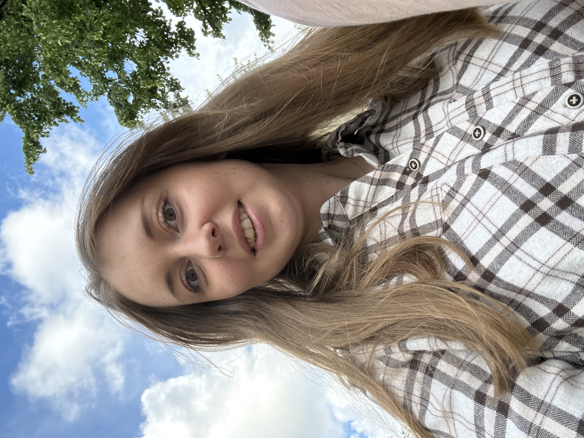 This media file is a close-up portrait of a young woman, rotated 90 degrees clockwise. The woman's face is positioned on the left side of the frame, with her body and shoulders extending towards the right. She has long, light brown hair that flows across the image. She is wearing a plaid shirt with a pattern of white and brown/beige squares and lines. The shirt features dark buttons, each with a white plus sign or cross symbol in the center.
The woman has a light complexion, green or hazel eyes, and appears to be smiling faintly. Her mouth is slightly open, showing some teeth.
The background suggests an outdoor setting. A bright blue sky with scattered white clouds occupies the lower portion of the rotated image, while green tree foliage is visible in the upper left corner. The lighting is bright and natural, indicating it is daytime. The unusual angle, with the sky at the bottom of the frame and trees at the top, suggests the photograph was taken from a low perspective, possibly as a selfie looking upwards, and then rotated.