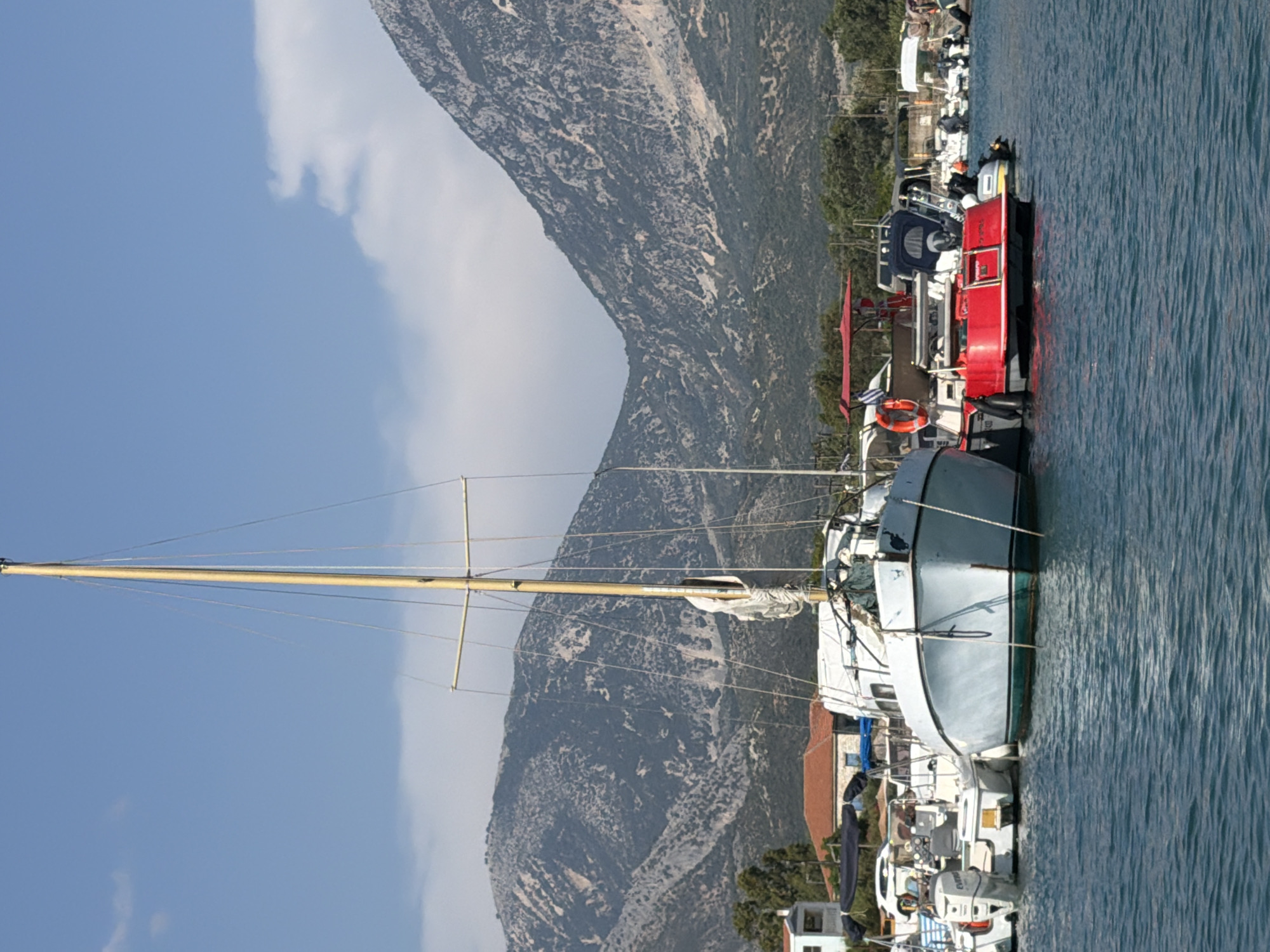 The image shows a marina with several boats docked in the Municipal Unit of Alyzia, Greece. In the foreground, a large sailboat with a tall mast is prominently featured, its hull painted light blue with a green stripe. Adjacent to it, a bright red speed boat is moored, with a life preserver visible on its side. Several other smaller boats, including some with outboard motors, are also docked in the marina. In the background, there are buildings with red-tiled roofs and trees, and behind them, a large, rugged mountain range under a clear blue sky with some scattered clouds. The water in the marina appears calm and blue. There are no people visible in the image.