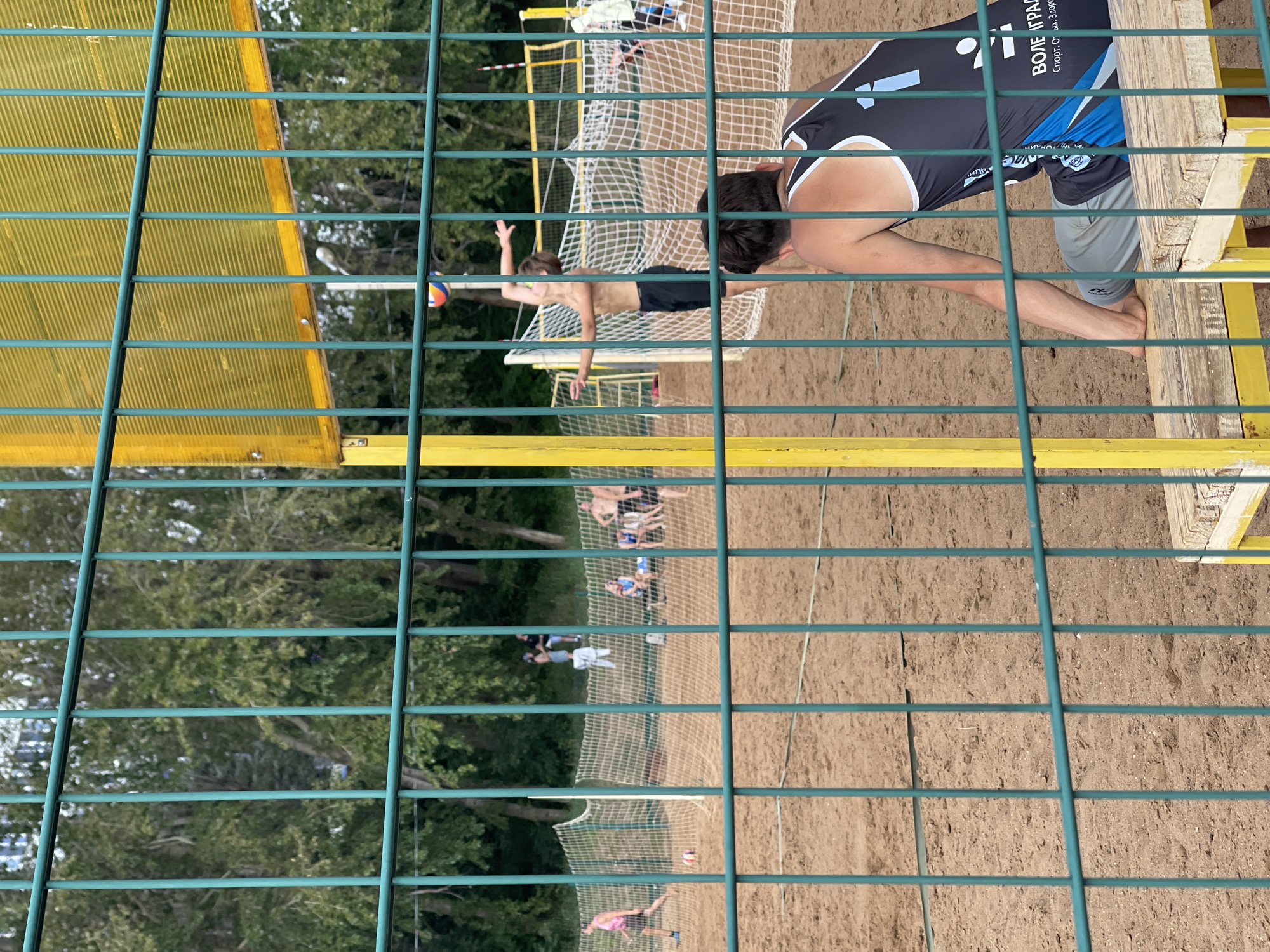 A group of people are playing beach volleyball on a sand court enclosed by a green metal fence. In the foreground, a person wearing a black and blue tank top sits on a wooden bench. In the background, several other individuals are actively engaged in the game, with one person in mid-air serving a volleyball. A yellow and green volleyball net is visible, as are trees in the distance. The scene appears to be an outdoor sports facility in Izhevsk, Russia.